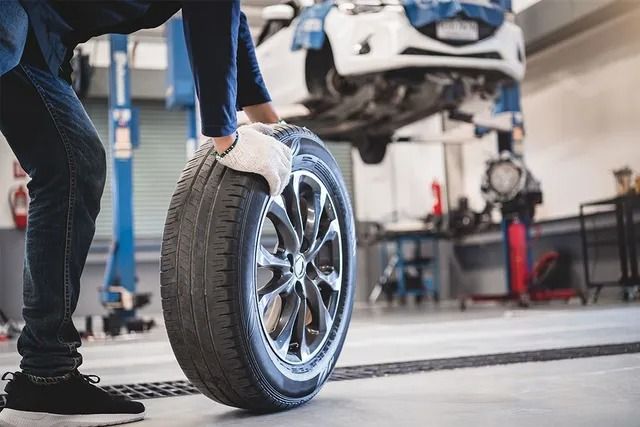 Mechanic in gloves rolling a tire in an auto repair shop with a car on a lift in the background.