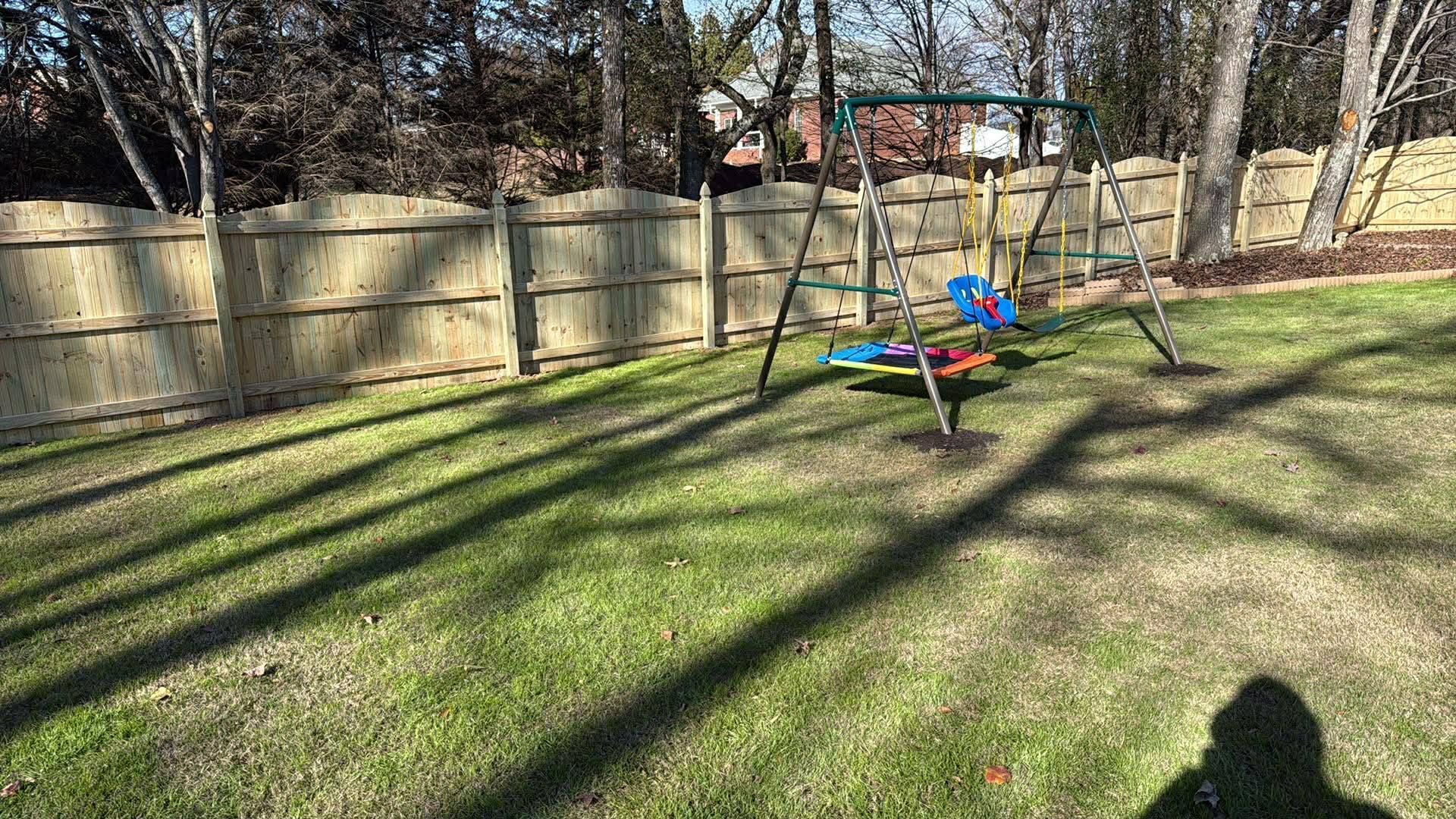 Backyard with a green lawn, wooden fence, trees, and a colorful playground toy in the center.