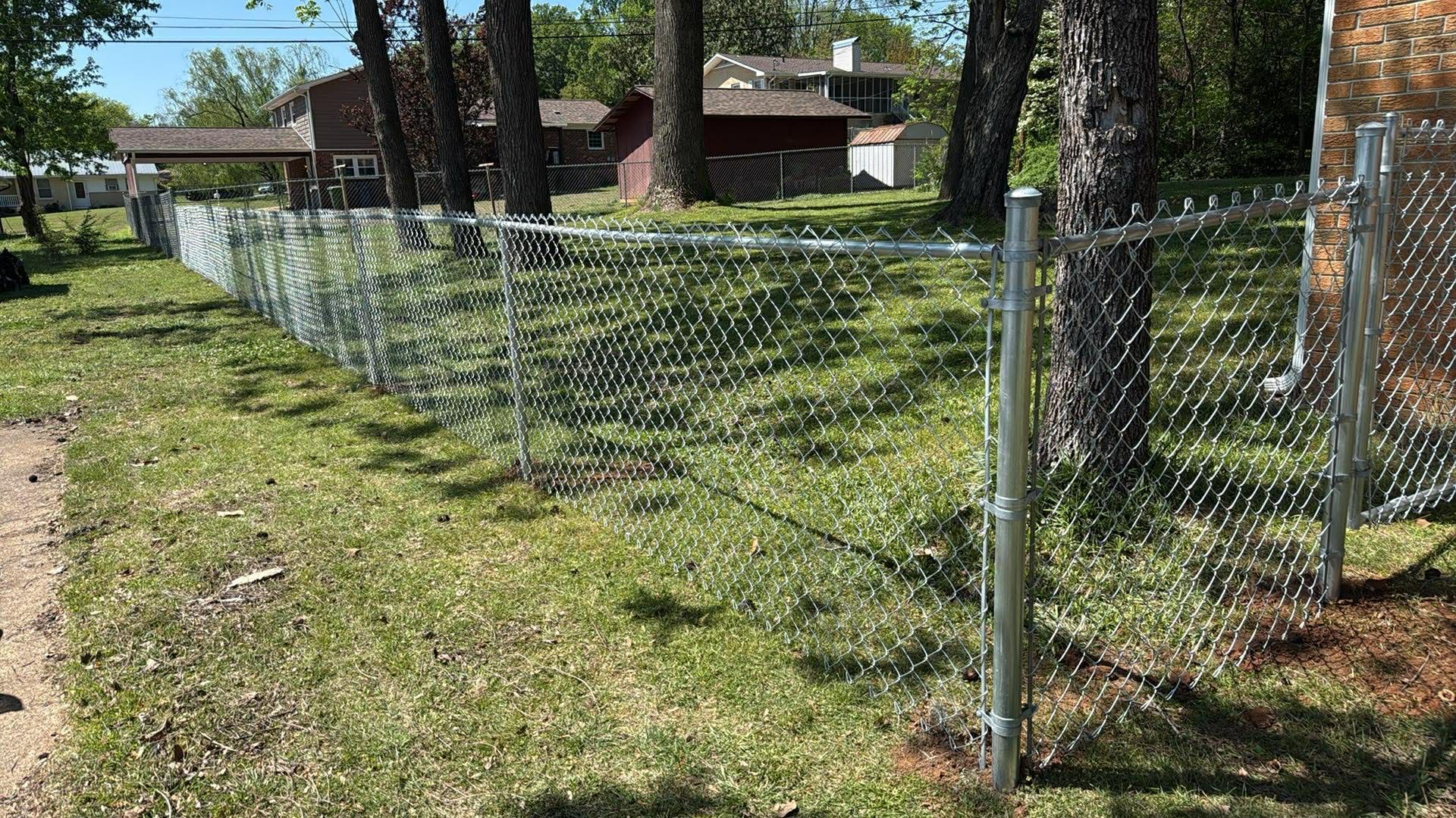 Chain-link fence along a grassy yard beside trees and a house on a sunny day.
