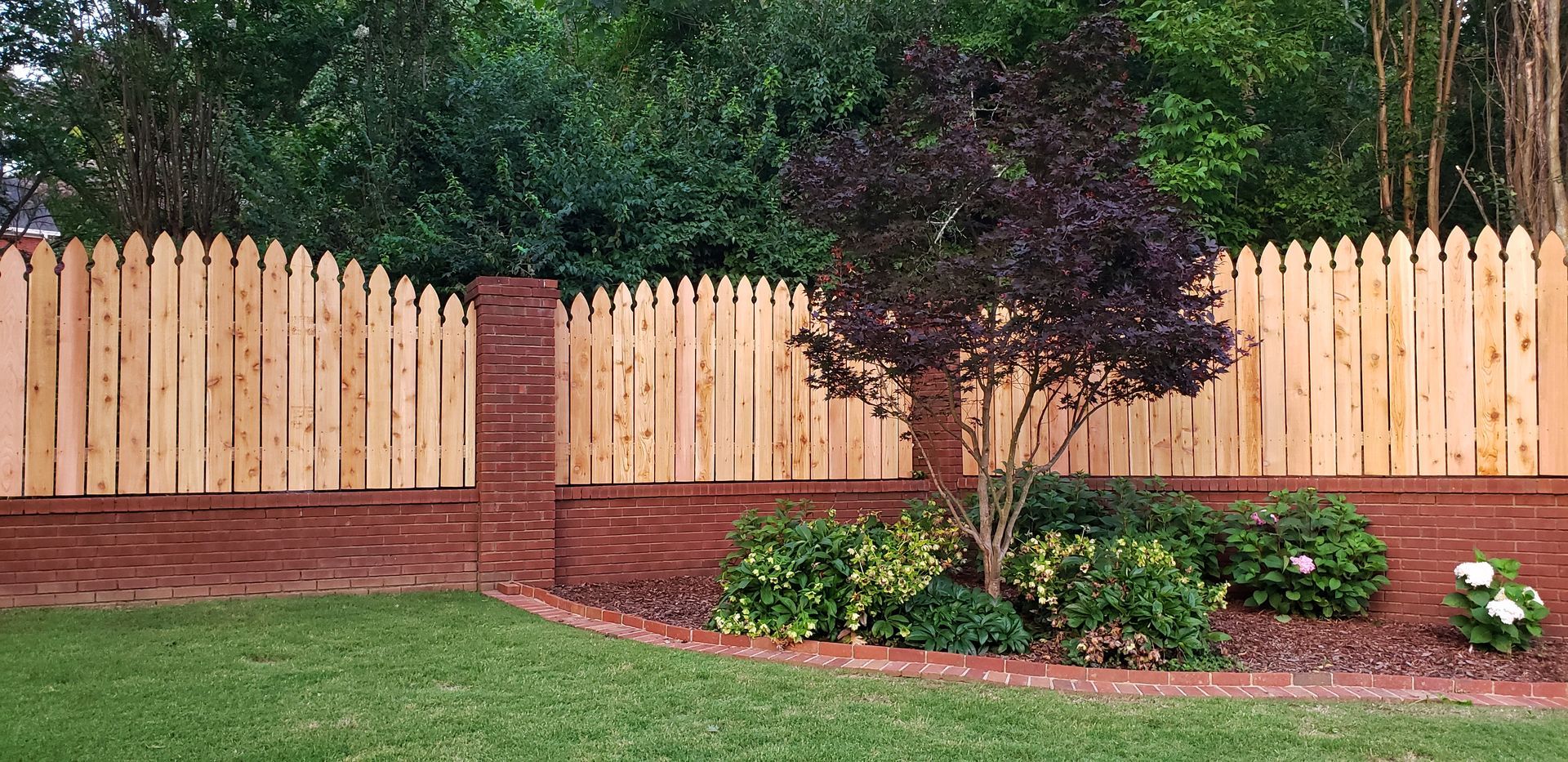 Wooden picket fence atop a red brick wall, a tree and bushes in front of a green yard.