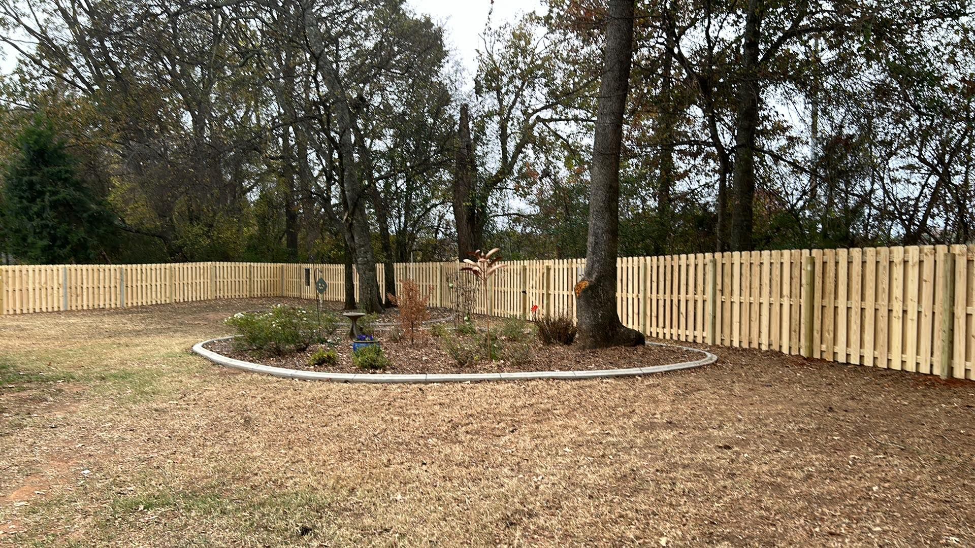 Wooden fence surrounds a yard with a circular garden and trees.