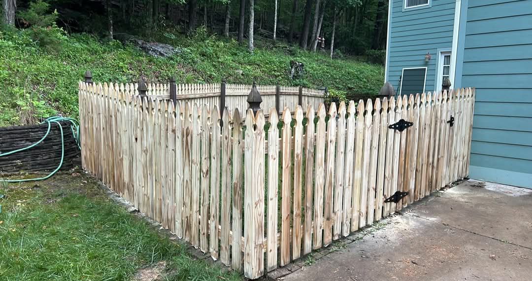 Wooden picket fence surrounds a corner of a building, near a grassy hill.