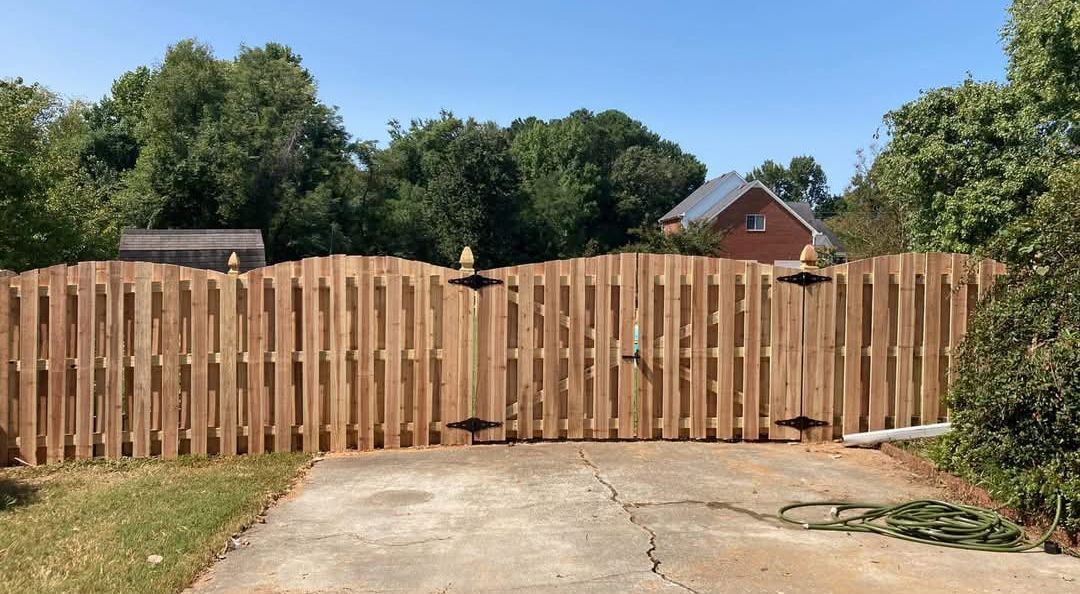 Wooden fence with a gate, trees in the background, and a driveway in front.