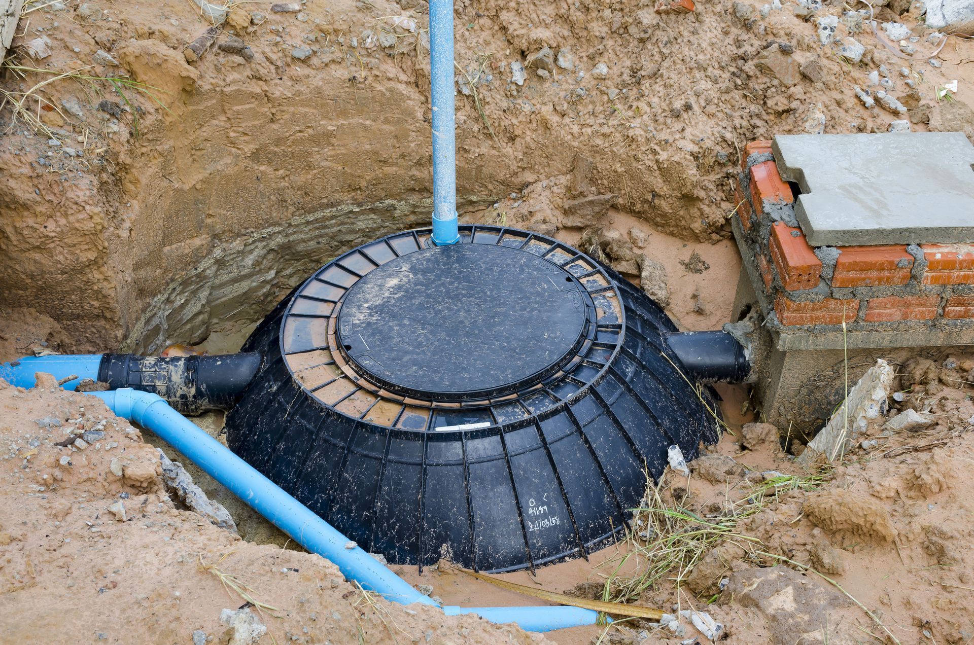 Black septic tank with access cover partially buried in sand with blue PVC pipes.