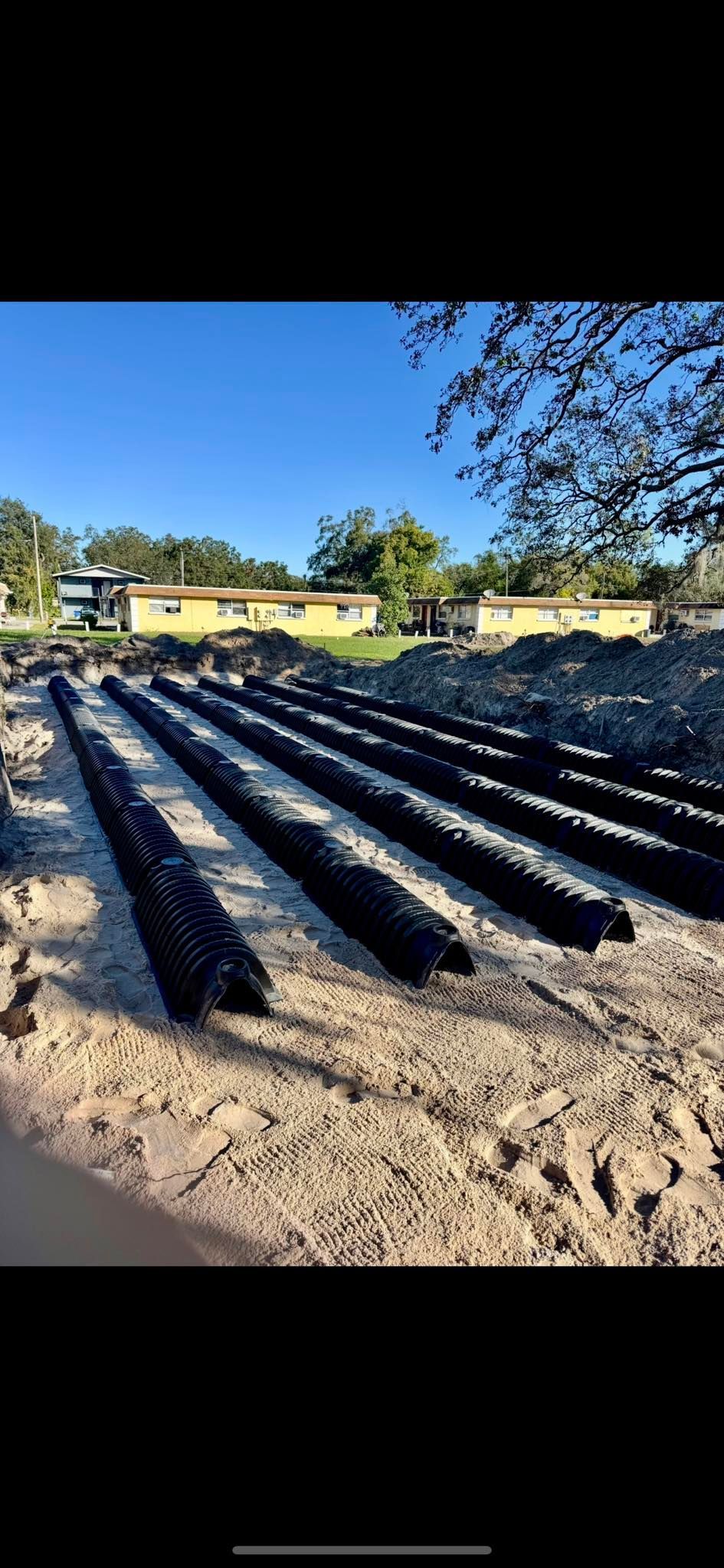 Rows of black drainage pipes laid in a trench, prepared for ground installation, set against a background of a blue sky and residential buildings.