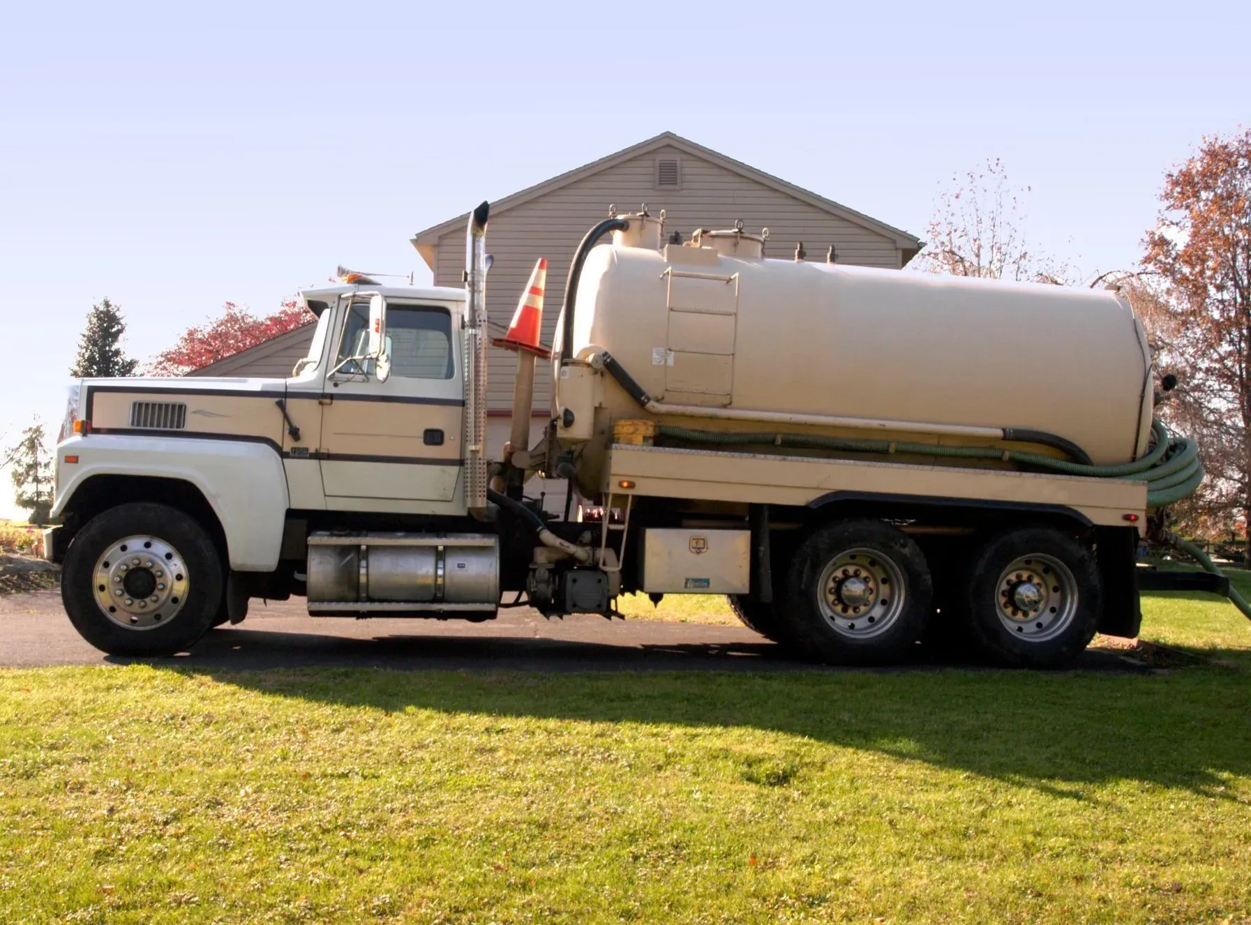 White and beige septic pumping truck on grass in front of a house.
