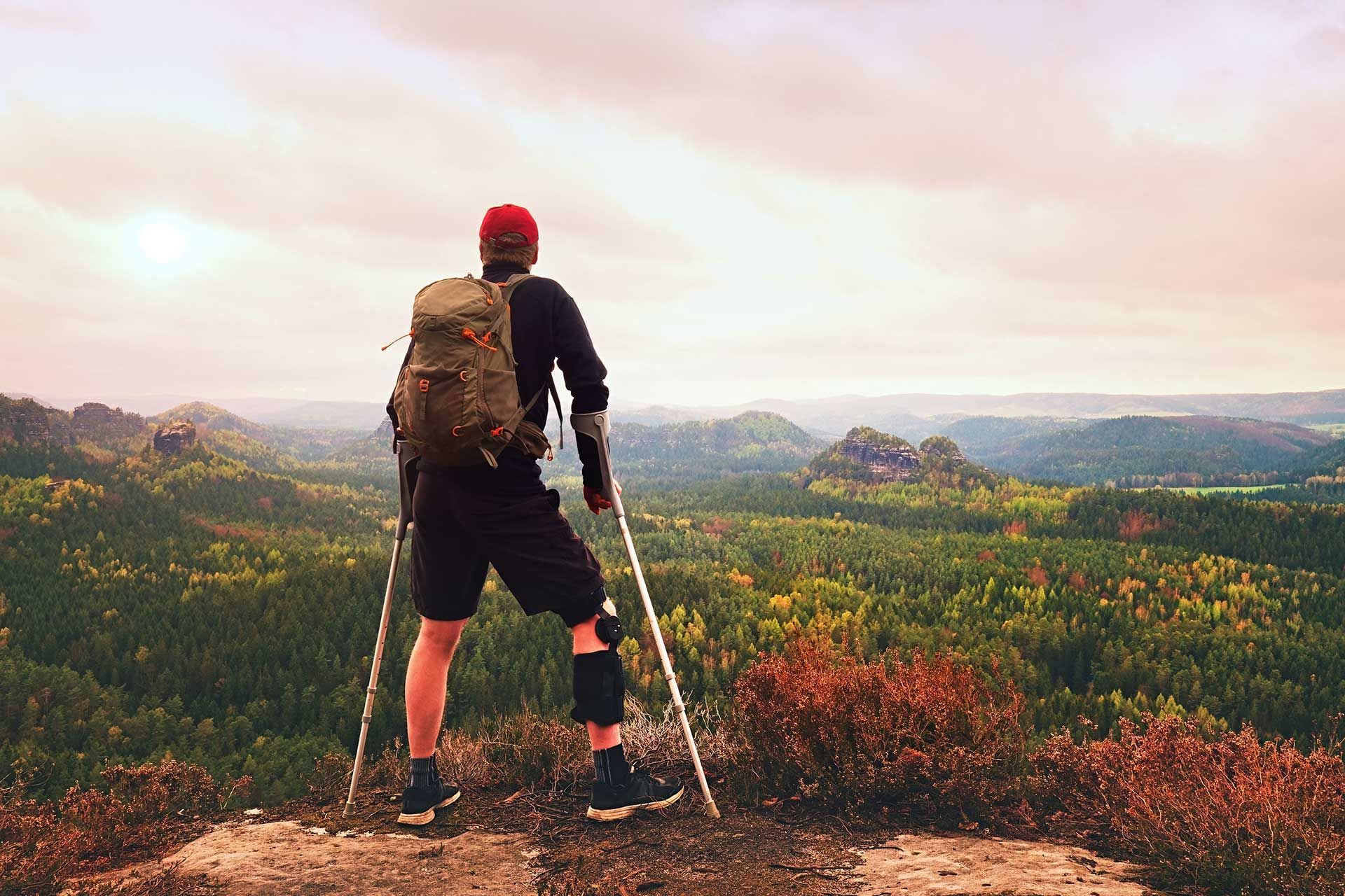 person with leg brace and crutches overlooking a forest from a mountain peak