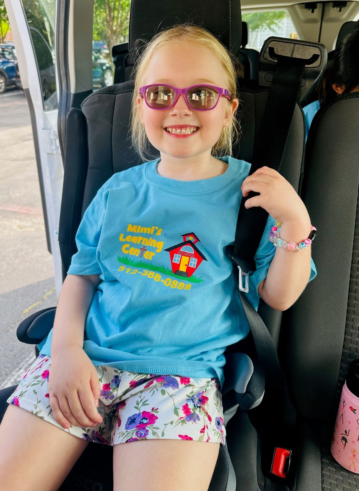 Smiling child in a car seat wearing pink sunglasses and a blue shirt, sitting in a vehicle's back seat