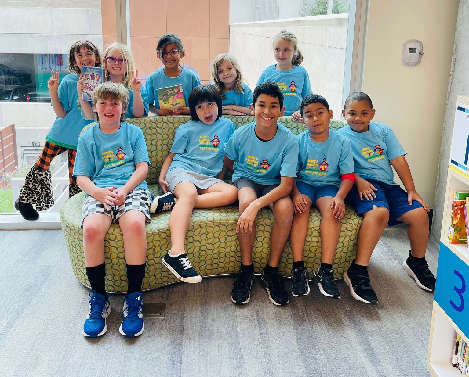 Group of children in matching blue shirts sitting on a bench in a bright indoor hallway