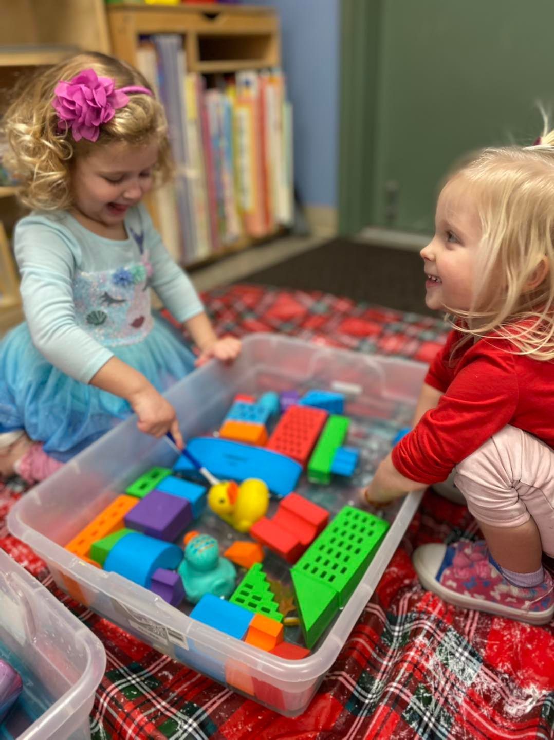 Two children play with colorful blocks in a classroom on a plaid blanket.