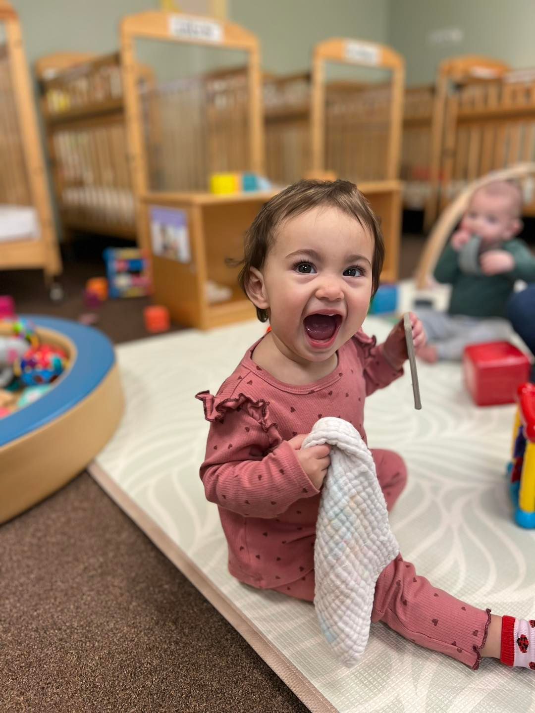 Smiling toddler in pink outfit holding a cloth, sitting on a rug in a playroom
