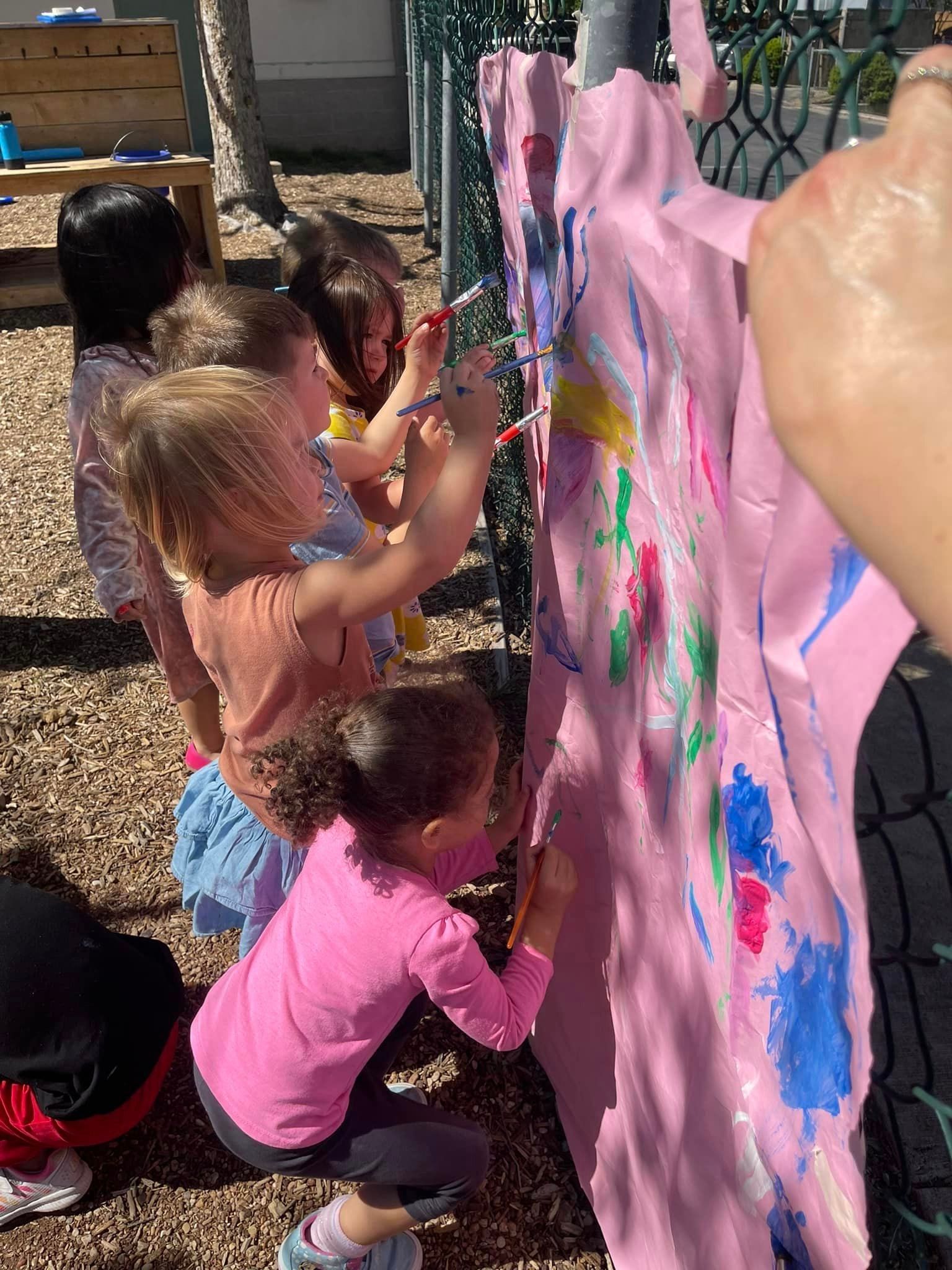 Children painting on a pink outdoor mural board with colorful handprints and brushstrokes
