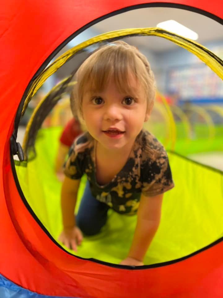Child crawling through a red tunnel in a colorful indoor play area, looking at the camera.