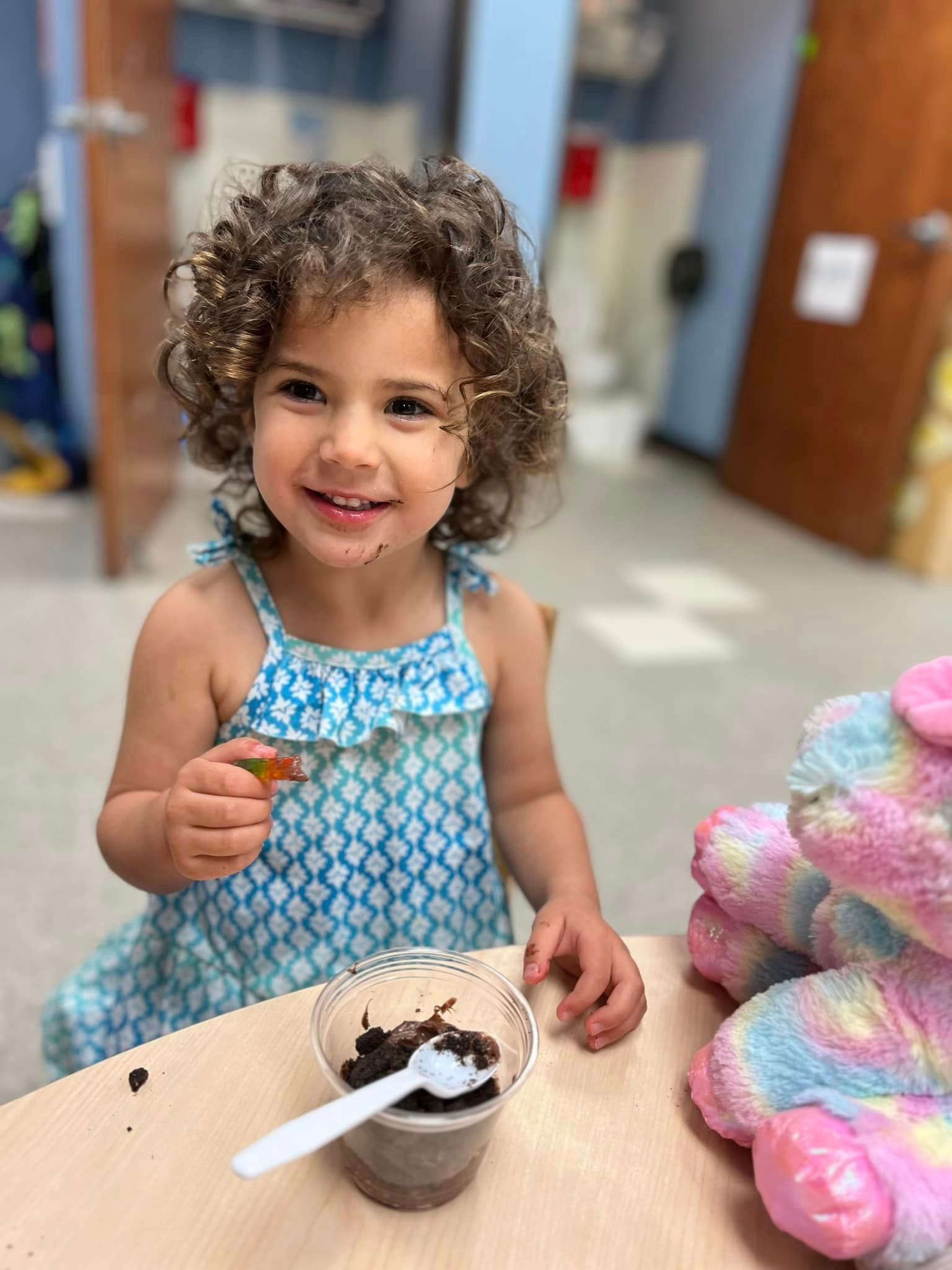 Smiling child in a blue dress holding a spoon beside a cup of dessert at a table