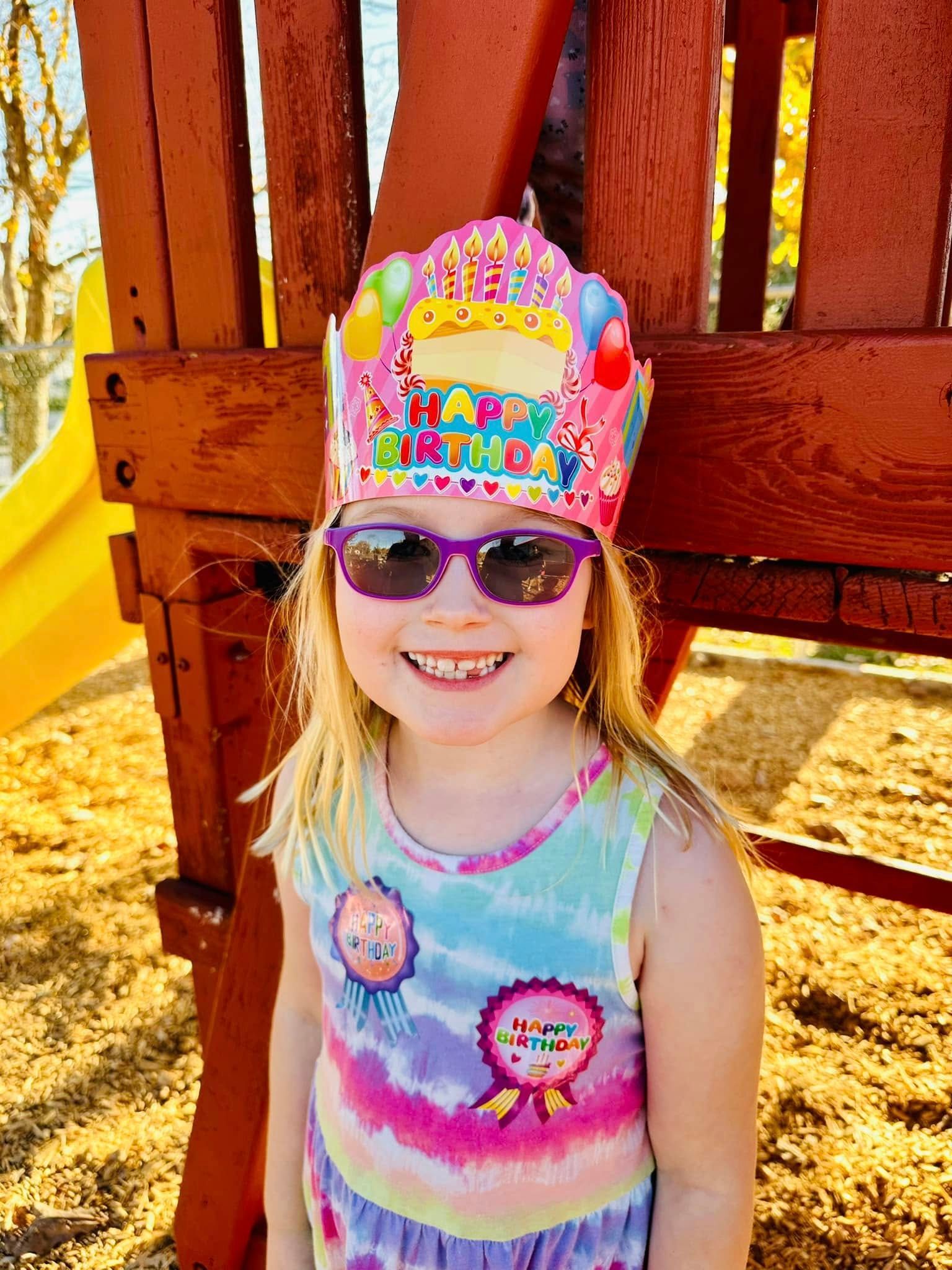 Child smiling in sunglasses and a colorful crown, standing by a wooden play structure and slide.