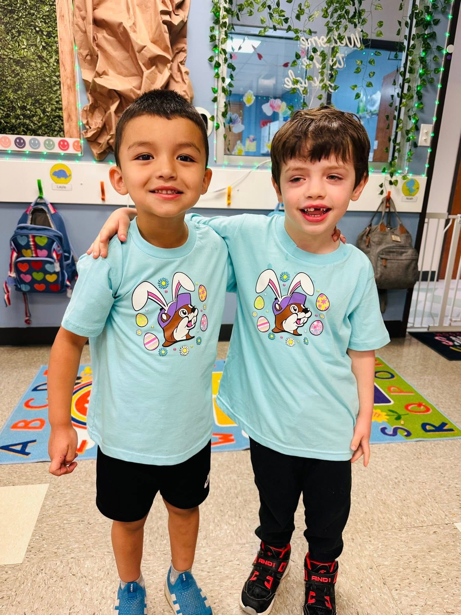Two children standing together in a classroom, smiling in matching mint green shirts.