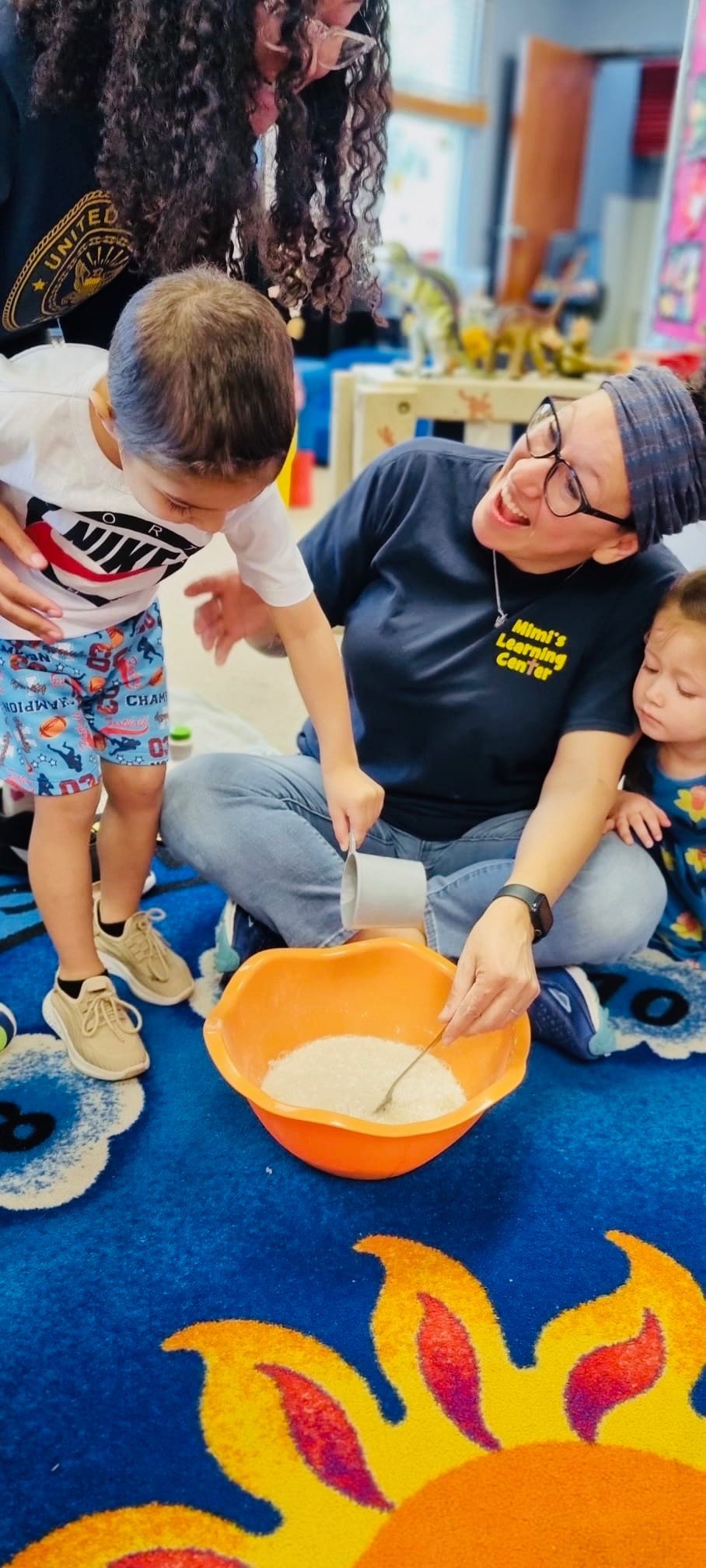 Three people kneel around a bowl of batter on a blue sun-patterned rug in a classroom.