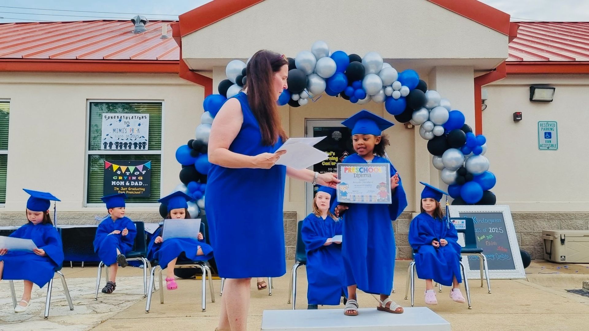 Graduates in blue caps and gowns on a decorated school stage with a balloon arch, holding diplomas