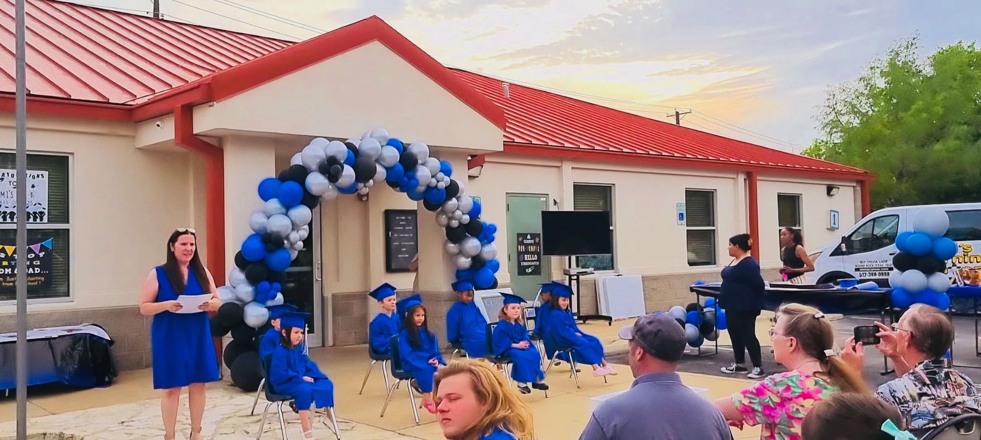 People seated outdoors at a blue-and-white balloon arch beside a white building with a red roof
