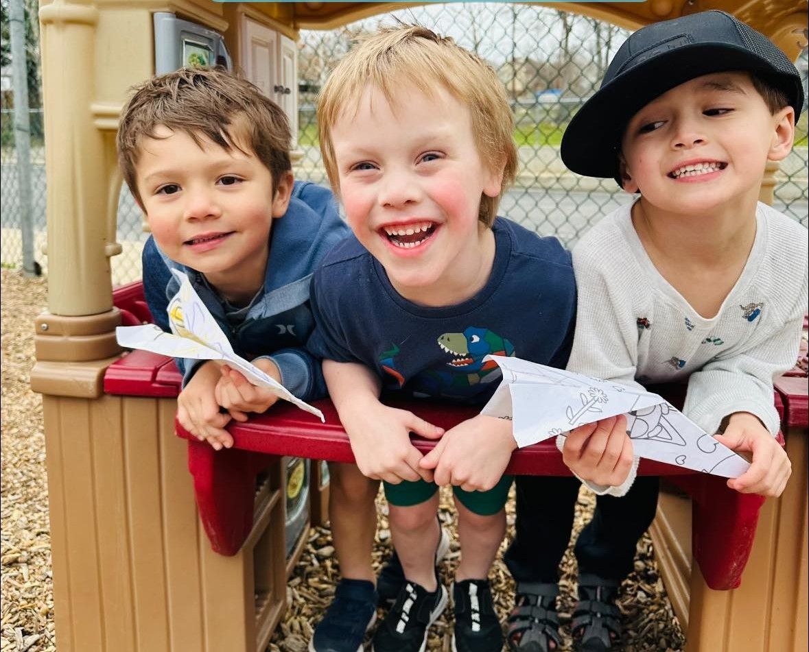 Three children leaning over a playground railing, smiling and holding paper airplanes.