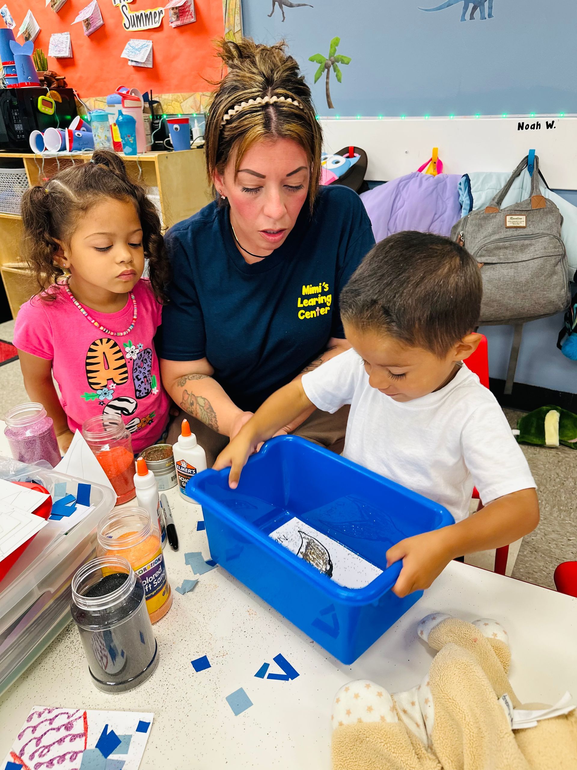 Adult and two children doing a craft activity at a table with a blue bin and paper scraps