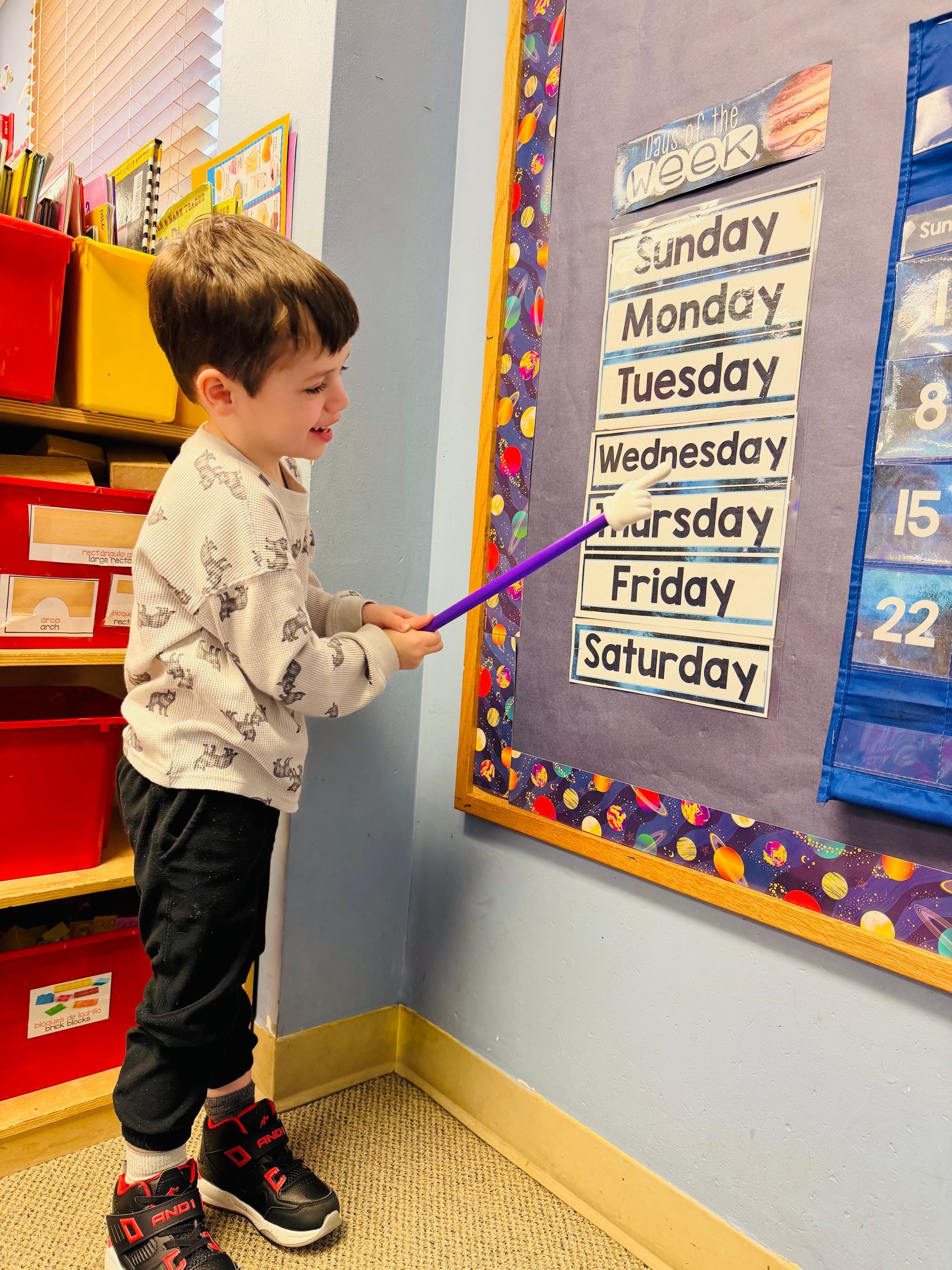 Child pointing at weekday names on a classroom wall chart with a purple pointer