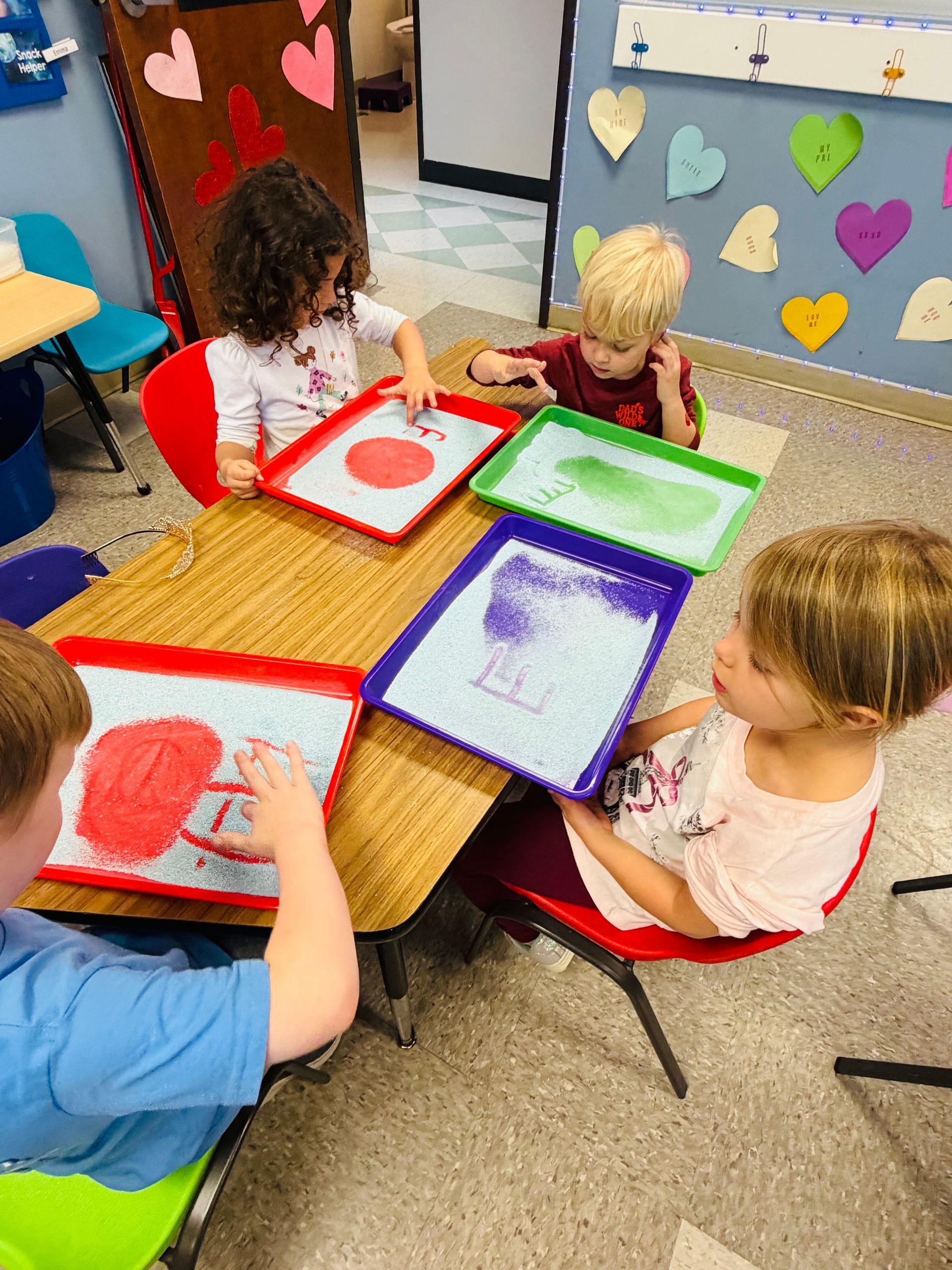Children painting at a table with colorful trays and paper in a classroom.