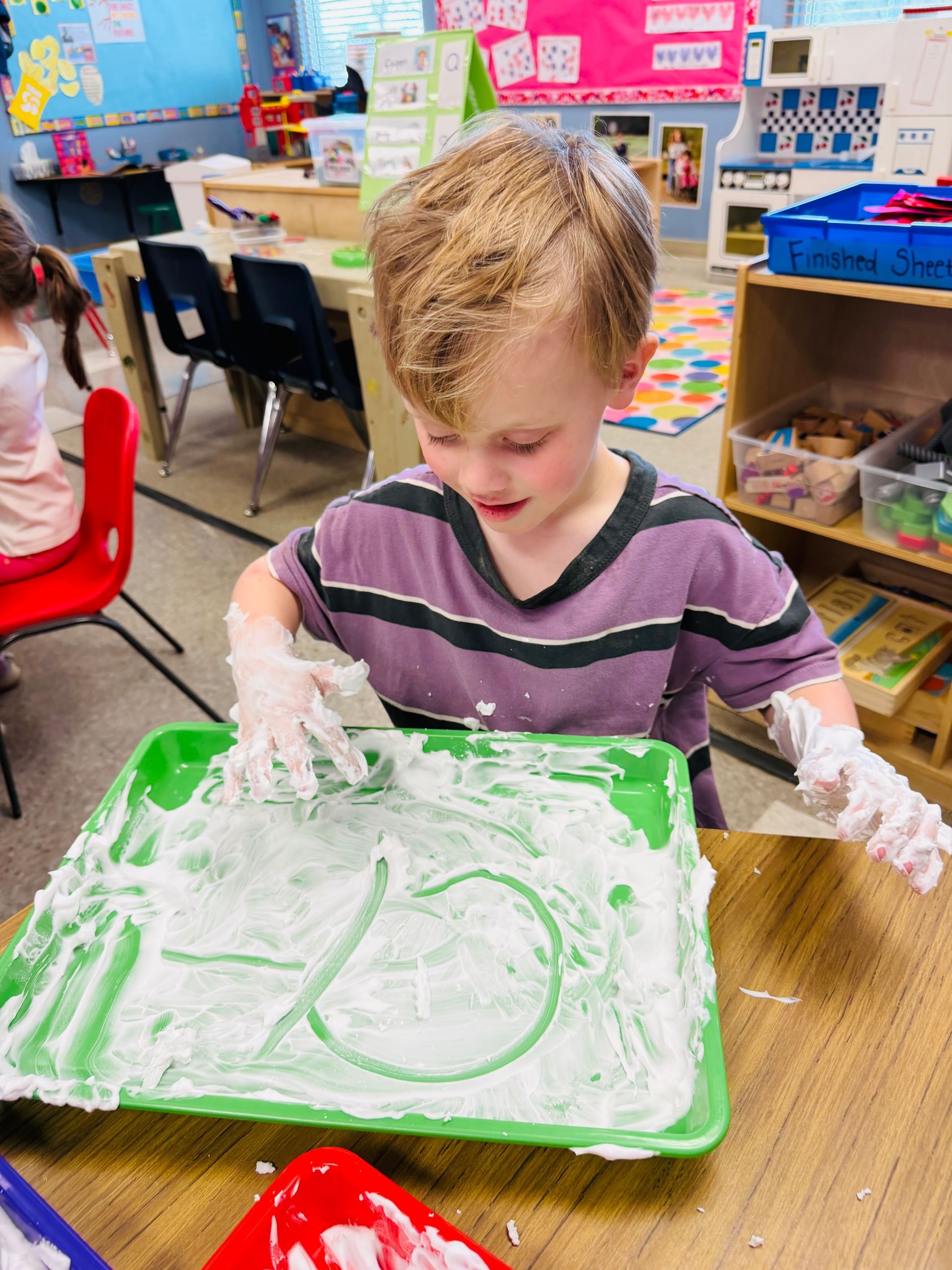 Child in a classroom spreads white foam on a green tray.