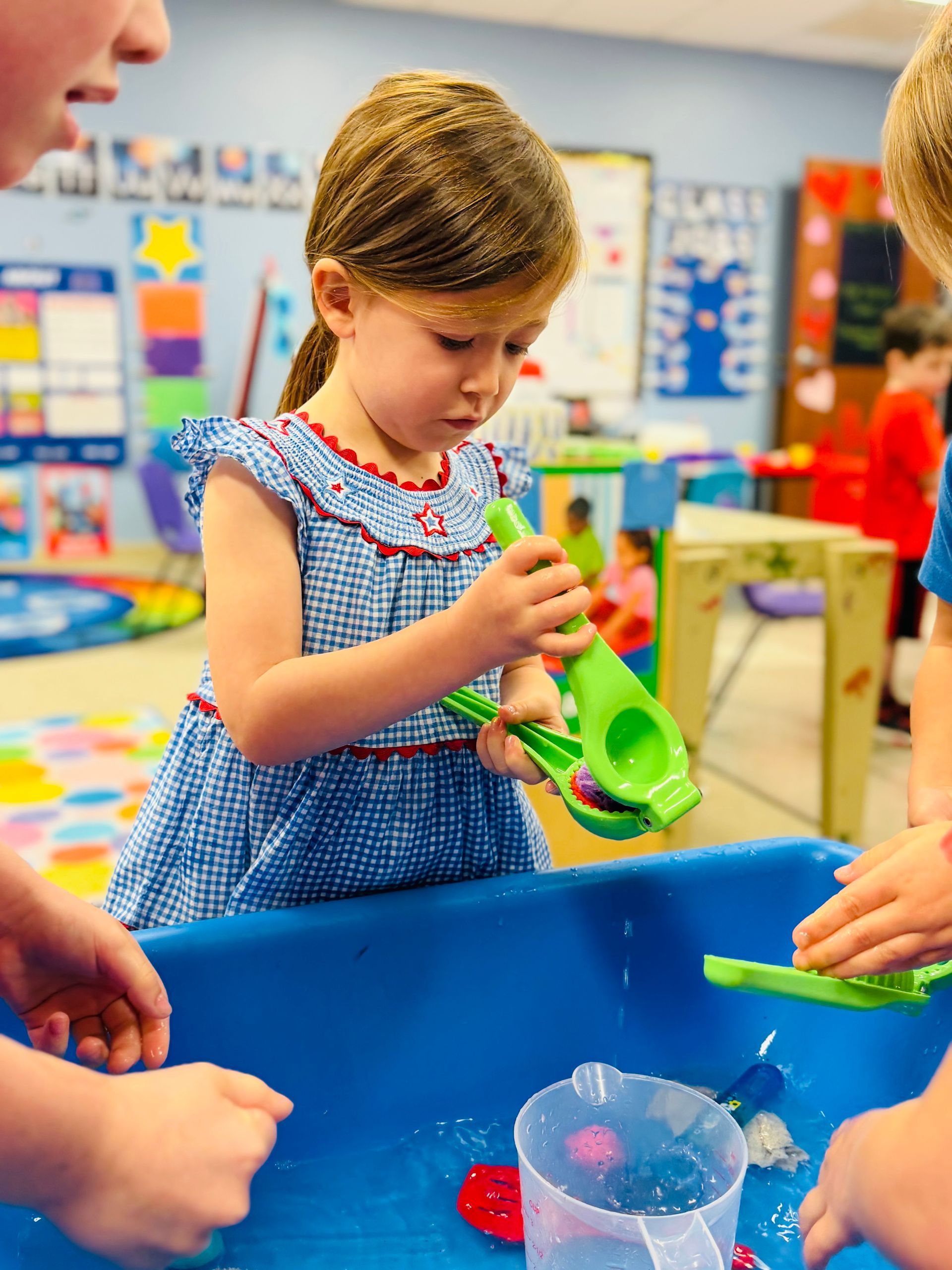Child pouring water into a green cup during a classroom activity with a blue bin and colorful toys.