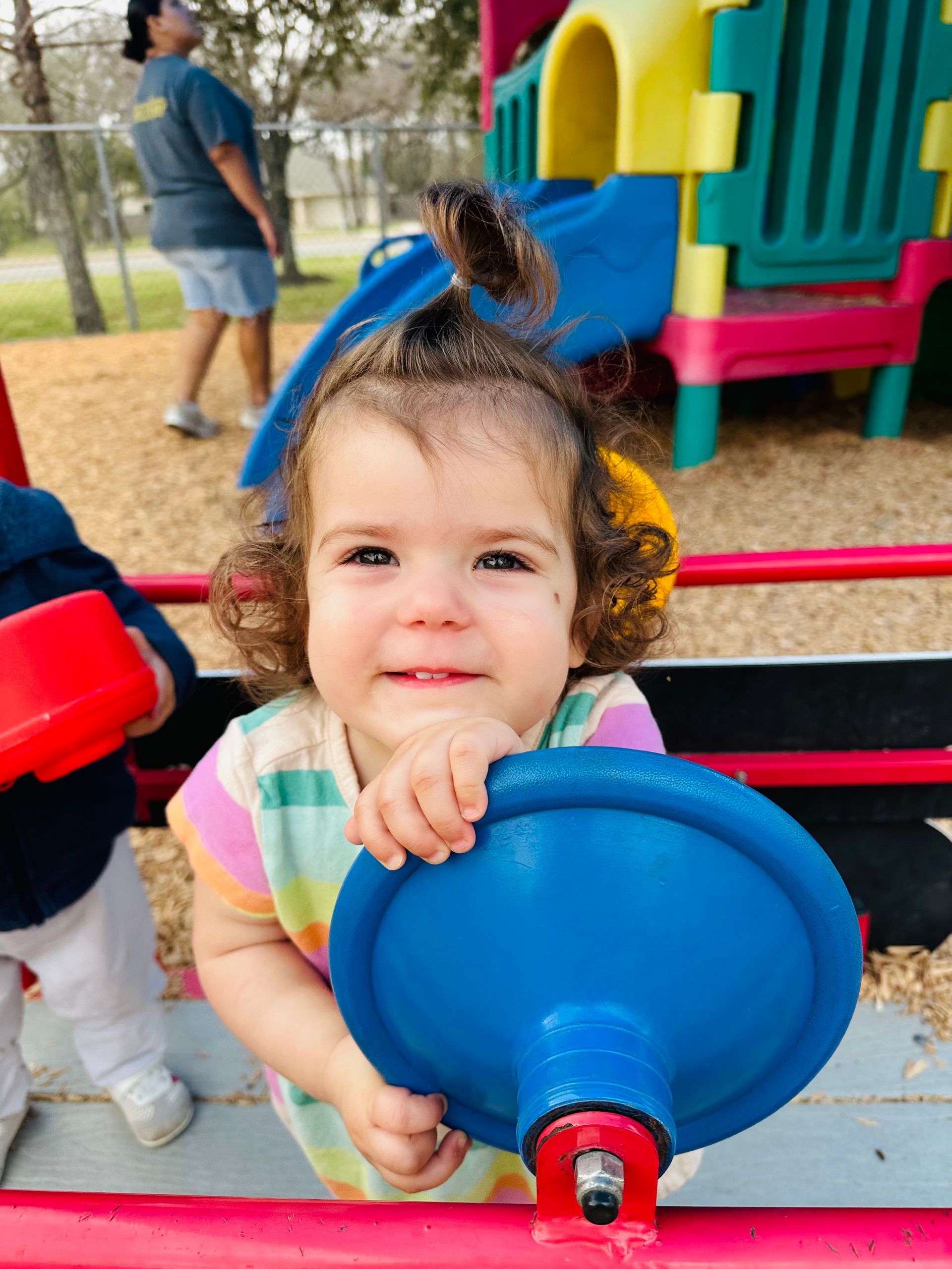 Smiling child at a playground, holding a blue spinning toy with colorful play equipment behind her