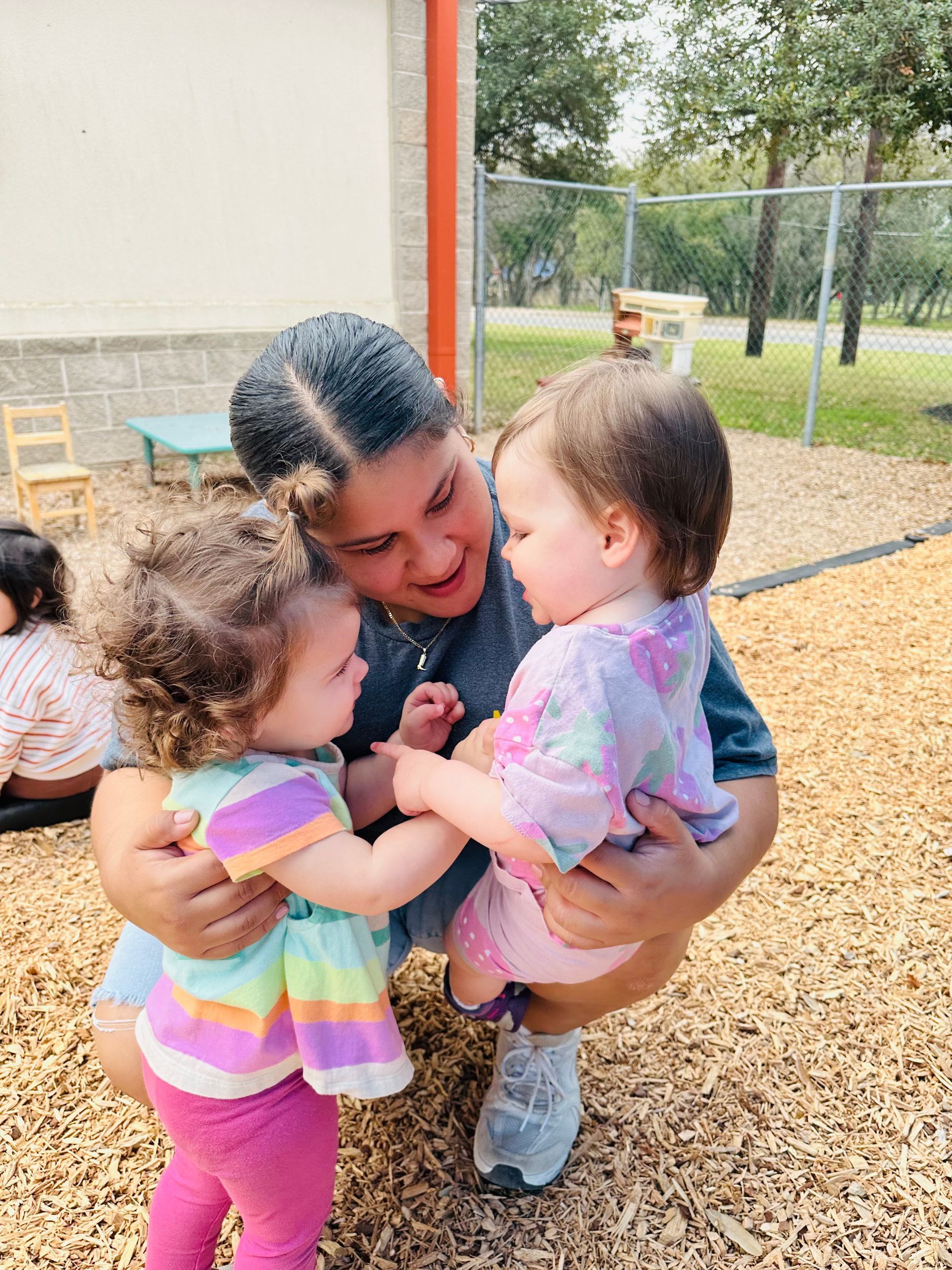 Three children hugging outdoors near a building, with a fence and trees in the background.