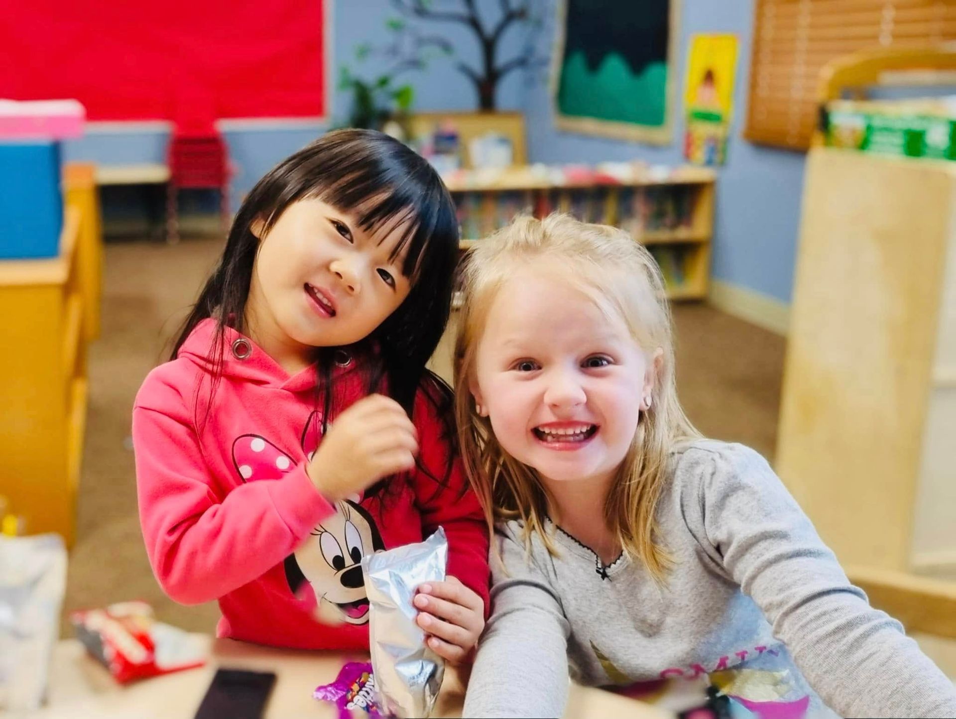 Two smiling children sitting together in a colorful playroom, holding a snack package.