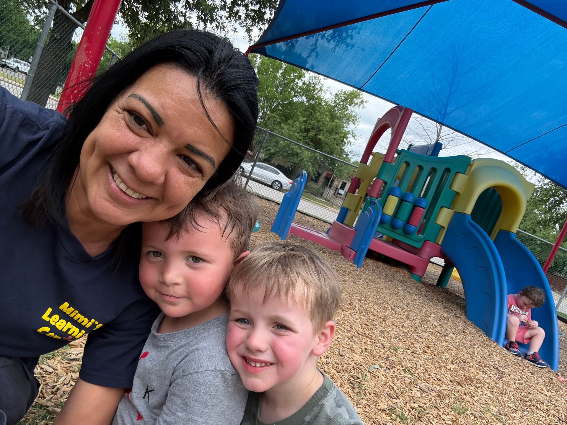 Three smiling people posing at a playground with colorful play structures and a blue shade canopy.