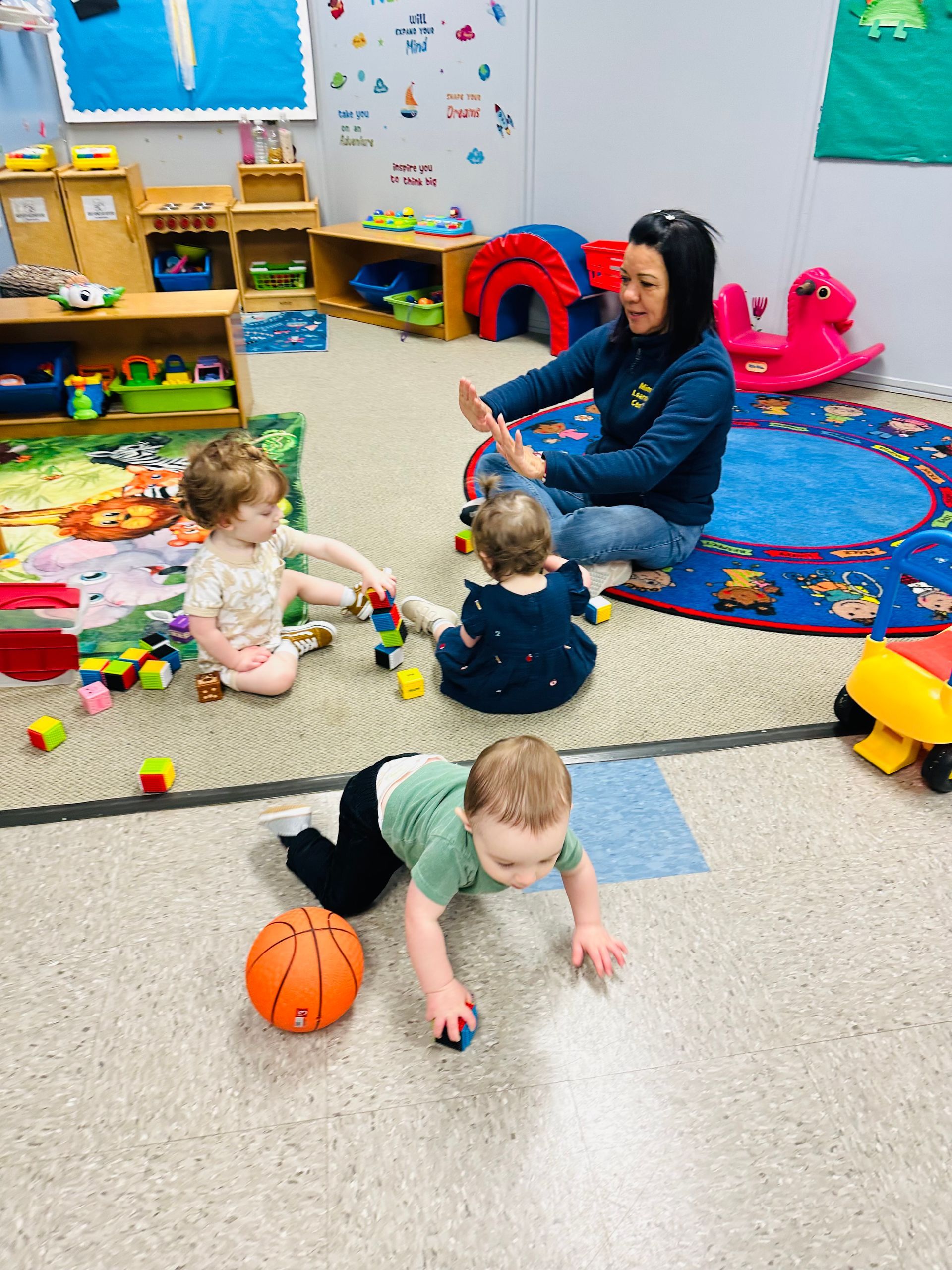 Toddlers playing on a daycare rug while a caregiver sits nearby among toys