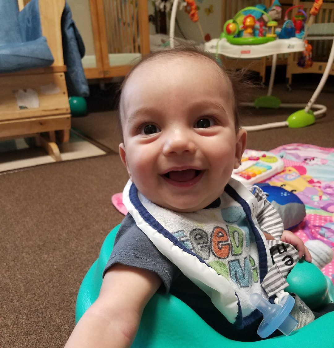 Smiling baby sitting on a play mat, reaching toward the camera in a colorful playroom.