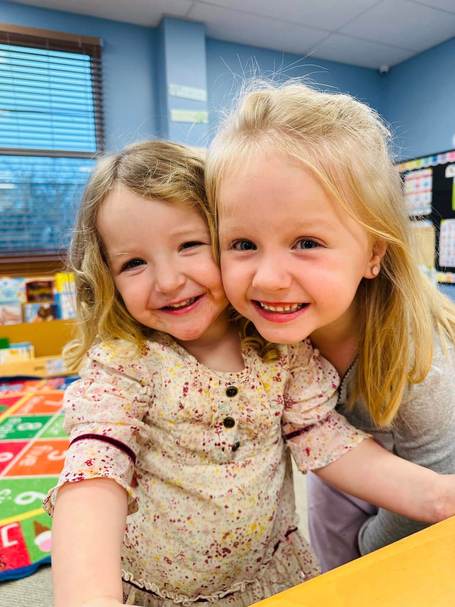 Two smiling children leaning close together in a colorful classroom.