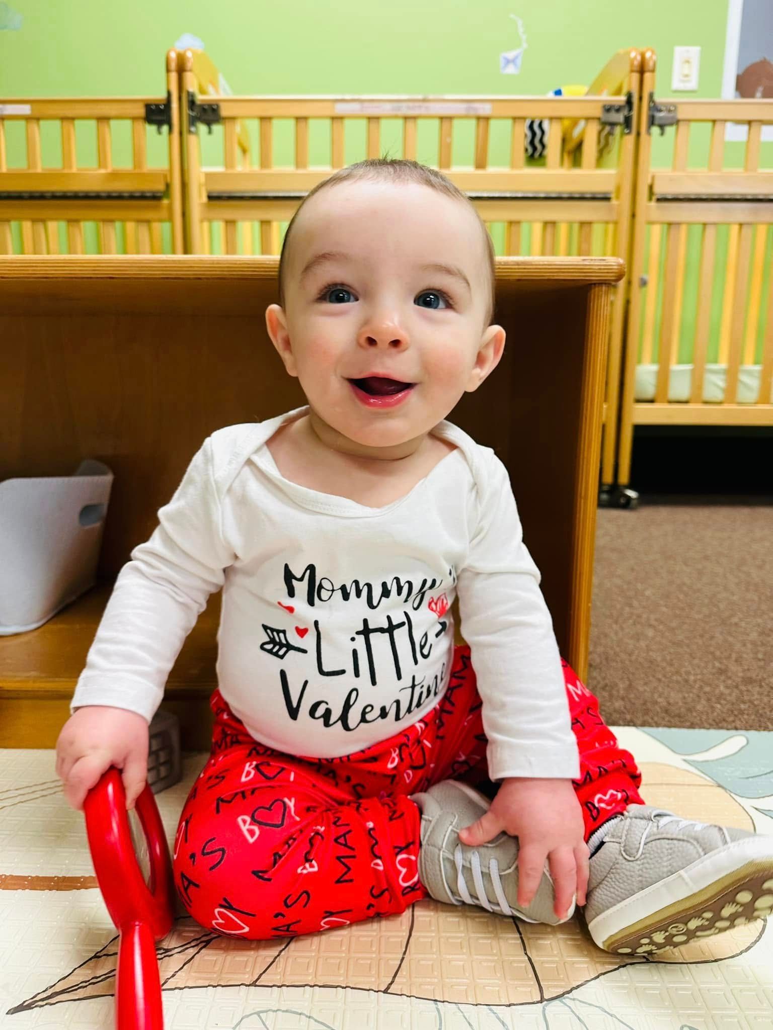 Baby sitting on a wooden floor in a nursery, smiling in a white shirt and red skirt with a gray shoe nearby.