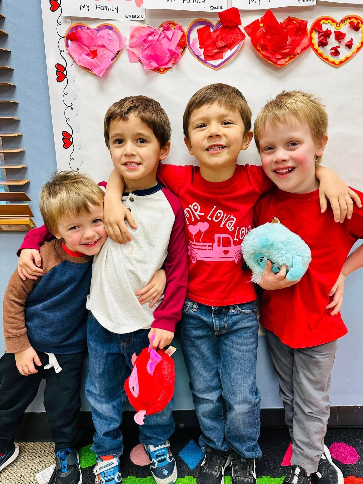 Four children standing arm-in-arm indoors, smiling and holding plush toys with pink heart decorations behind them