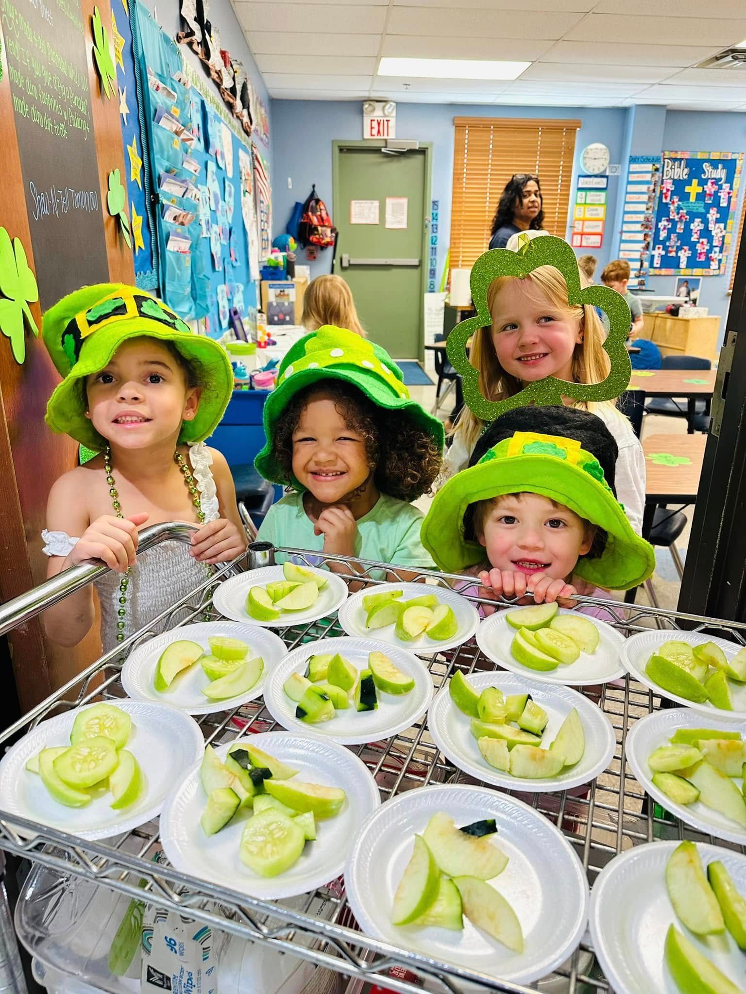 Children in green hats smiling behind a table of lettuce cups in a classroom cafeteria