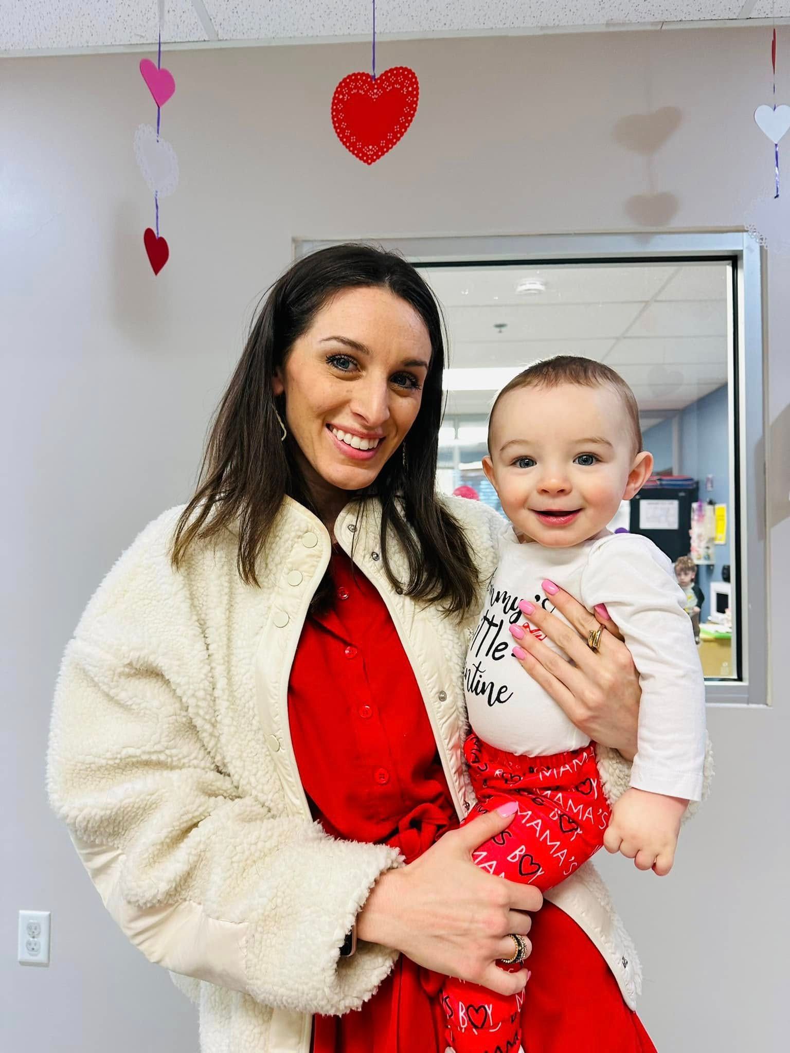 Woman holding a smiling baby in a white room with red heart decorations for Valentine’s Day.