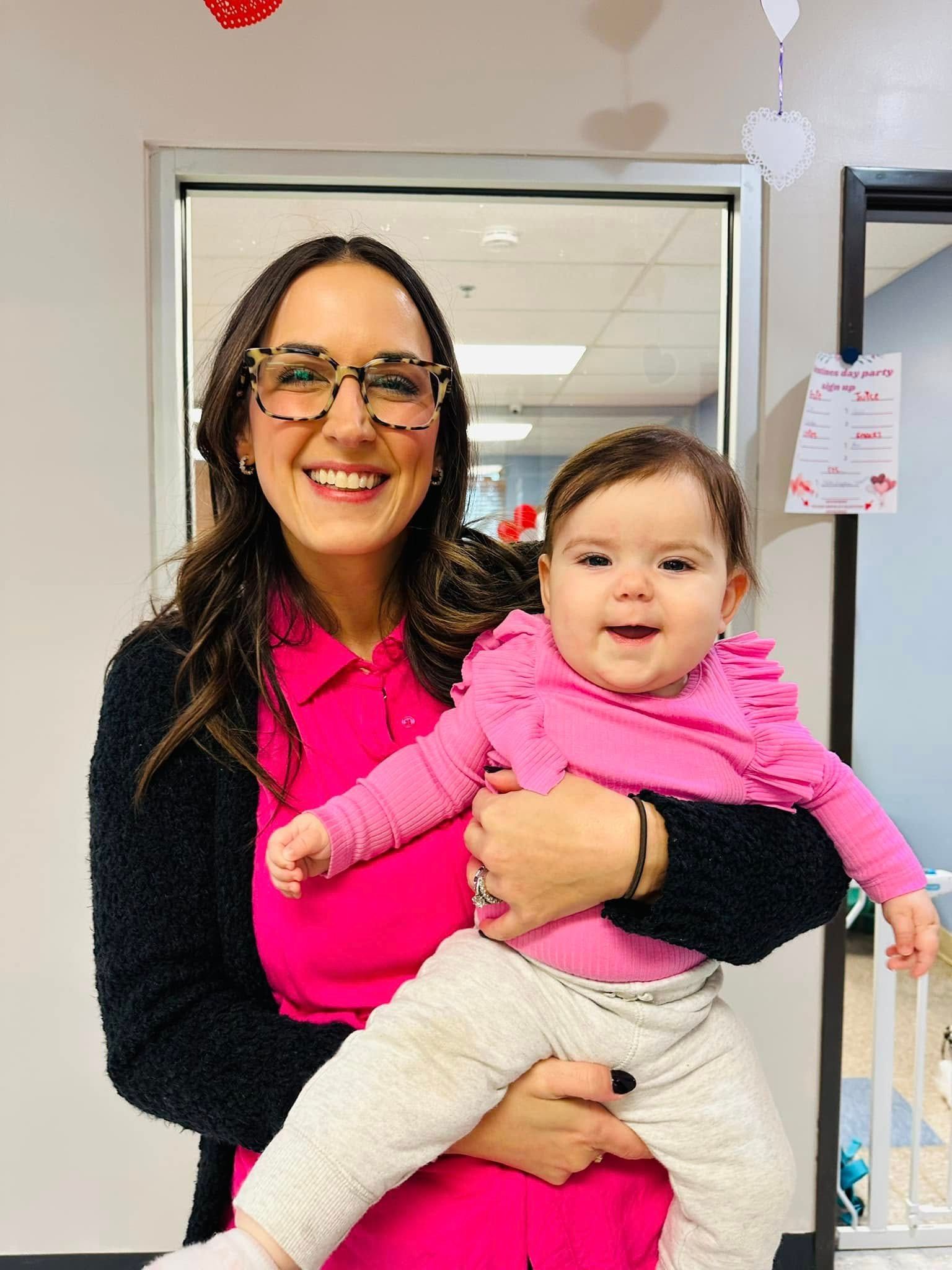 Smiling woman in pink holds a baby in pink and gray in an office hallway.