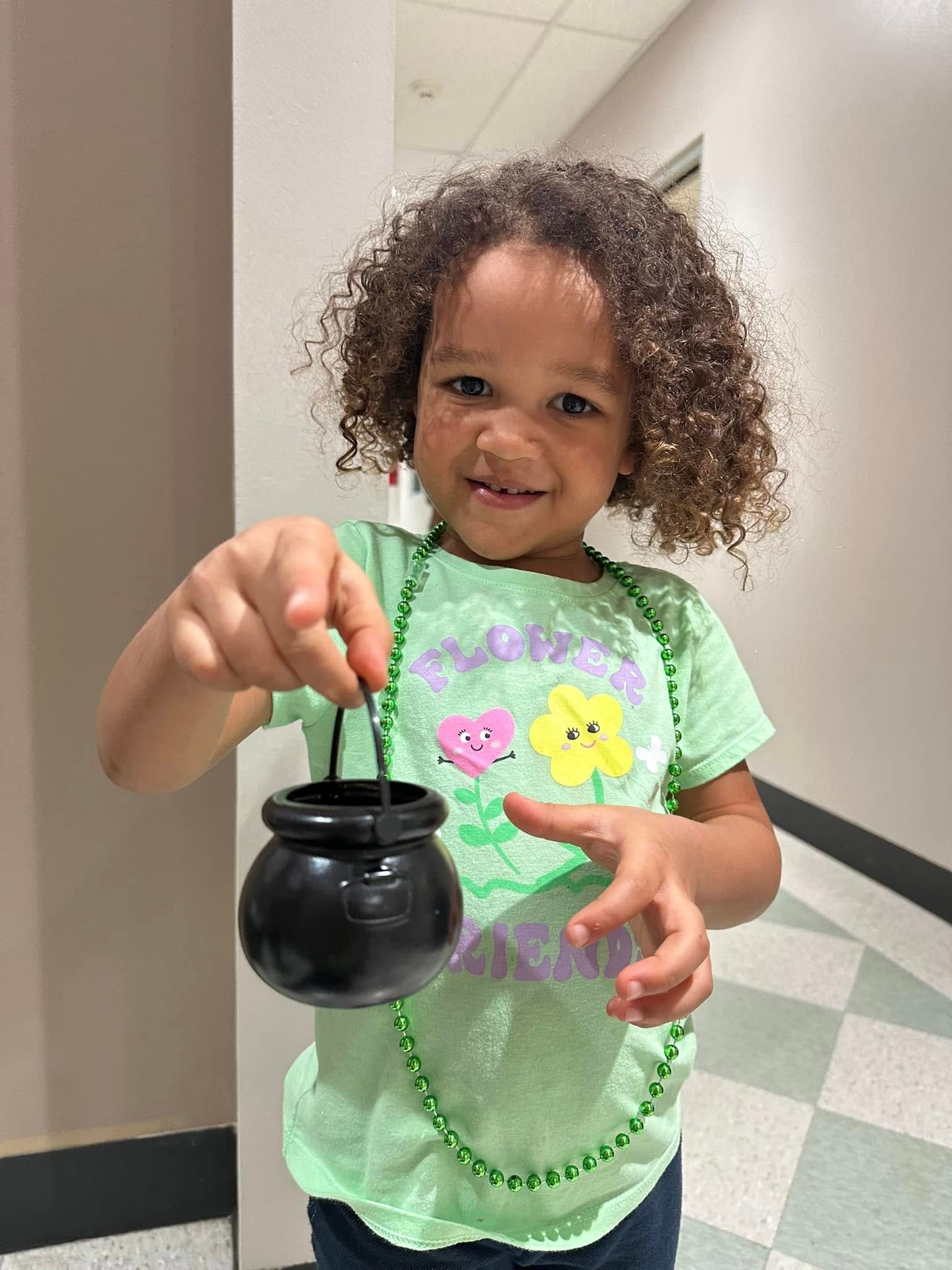 Child in a green shirt holding a black cauldron-shaped bucket in a hallway.