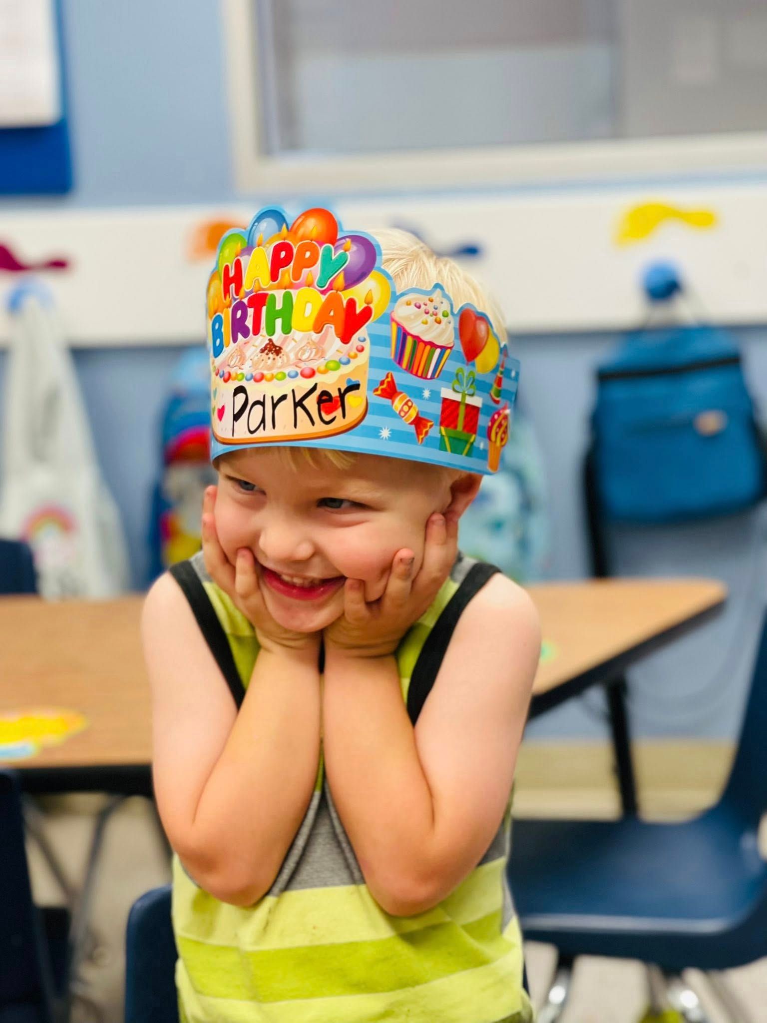 Smiling child wearing a colorful birthday hat and green shirt in a classroom, hands on cheeks.