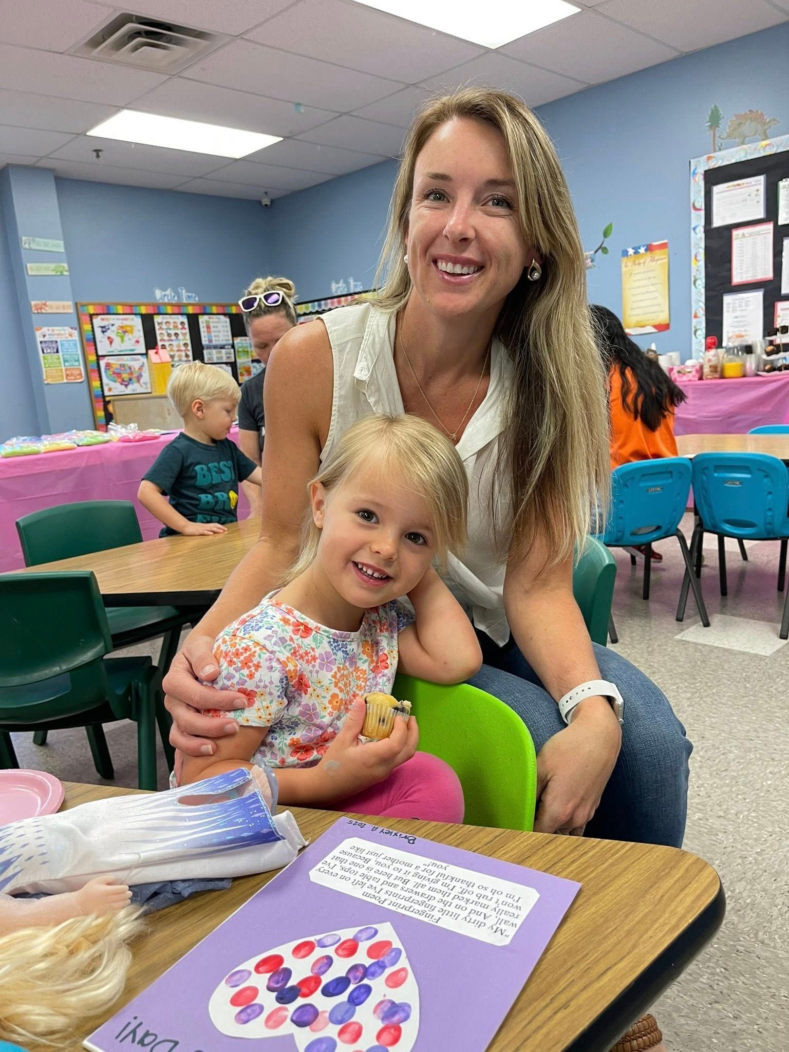 Woman and young child smiling in a classroom, seated at a table with craft supplies.