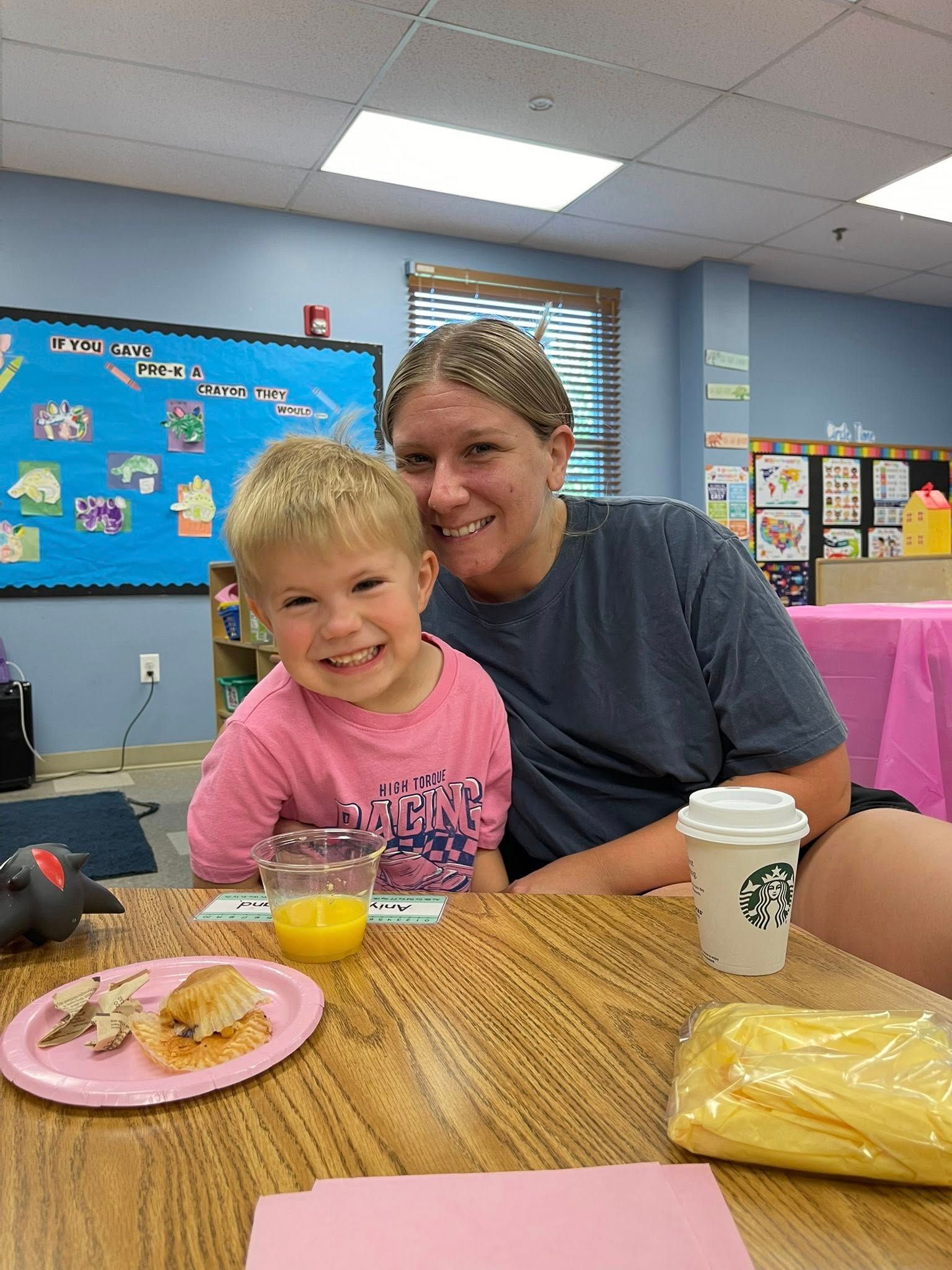 Smiling adult and child seated at a table in a classroom, with breakfast food, coffee, and juice.
