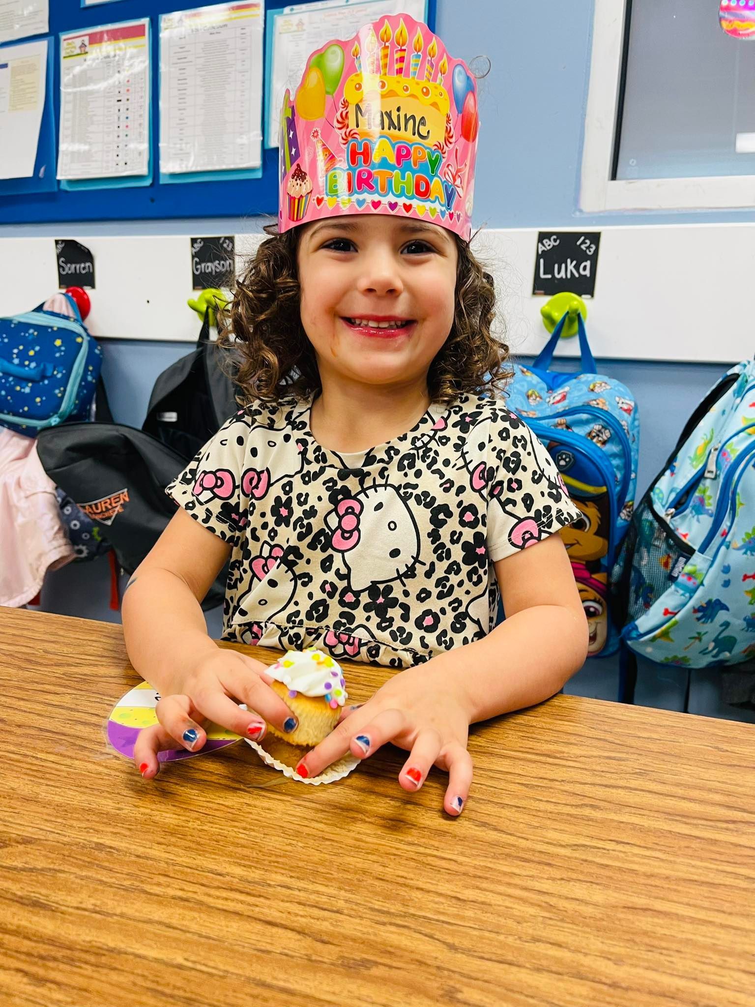 Child smiling at a classroom table, wearing a colorful birthday crown and holding a small toy.