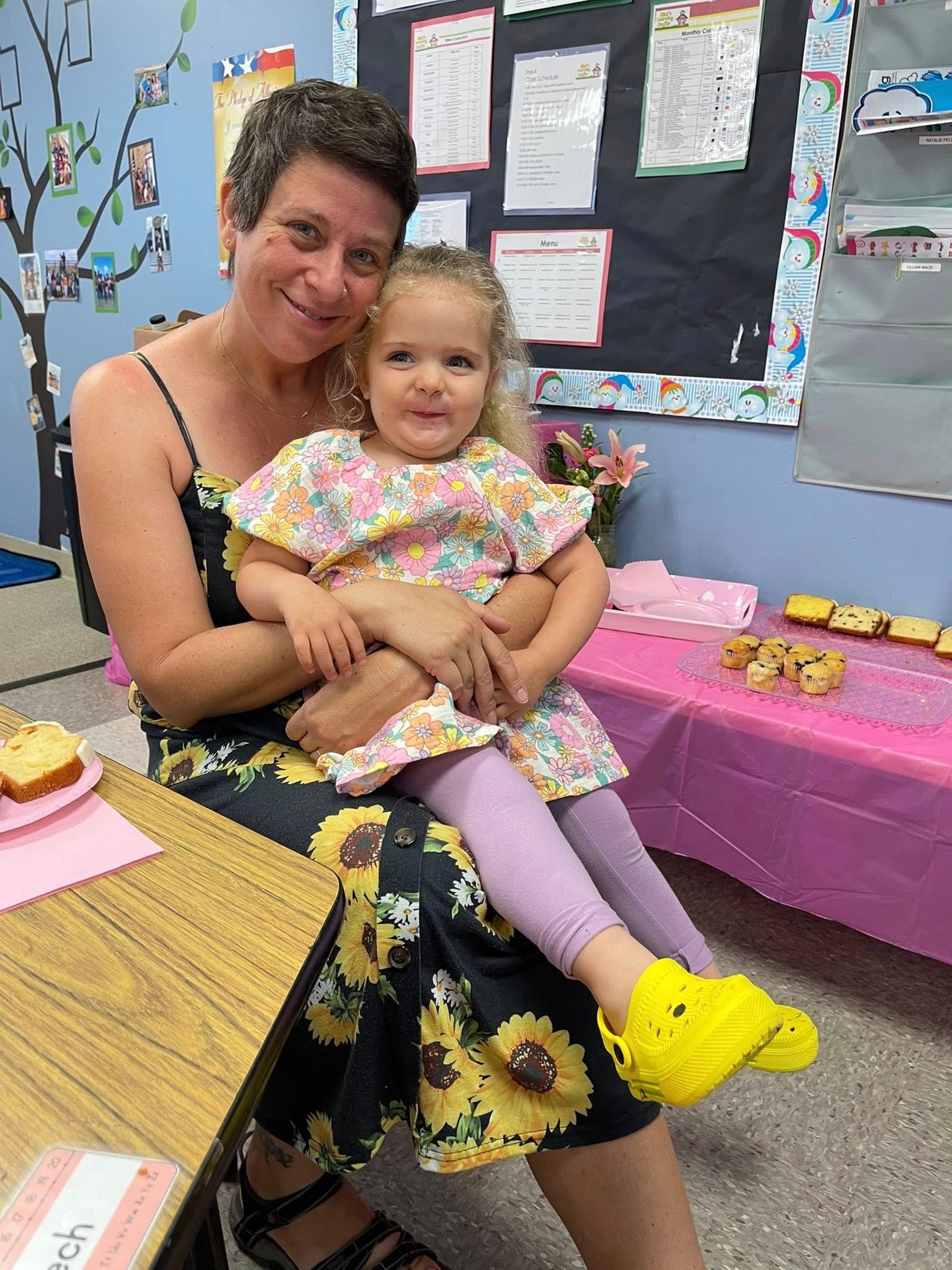 Woman holding a young child at a decorated event table with pink tablecloths and a bulletin board behind them
