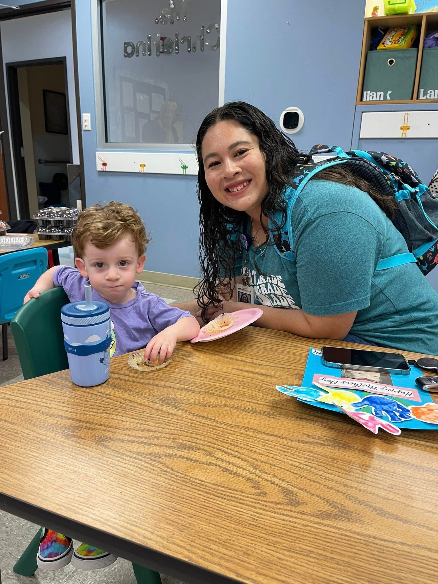 Adult and child seated at a table in a classroom, smiling with coloring materials and a water bottle.