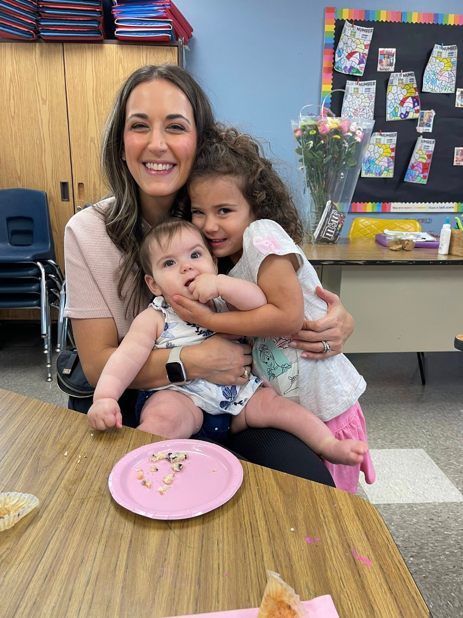 Woman with two children smiling at a classroom table, with a pink plate in front of them.