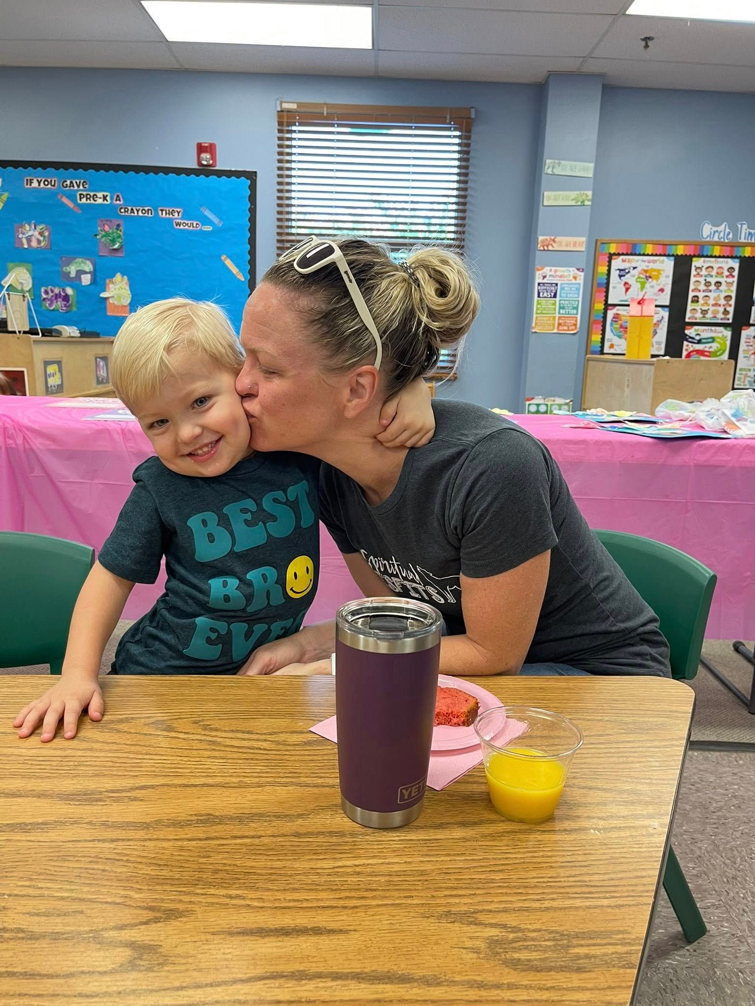 Child smiling while being kissed on the cheek at a table in a classroom, with a drink and small cup nearby